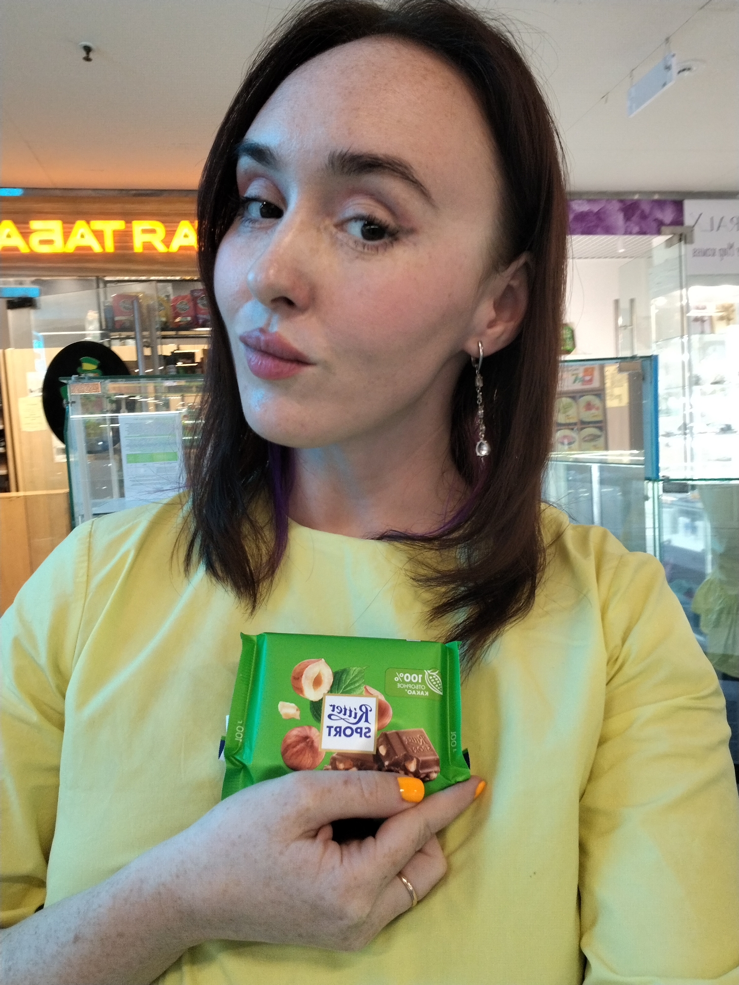 A woman with dark hair and freckles is holding a Ritter Sport chocolate bar. She is wearing a yellow shirt and has orange nail polish. The background appears to be a store with bright signage.