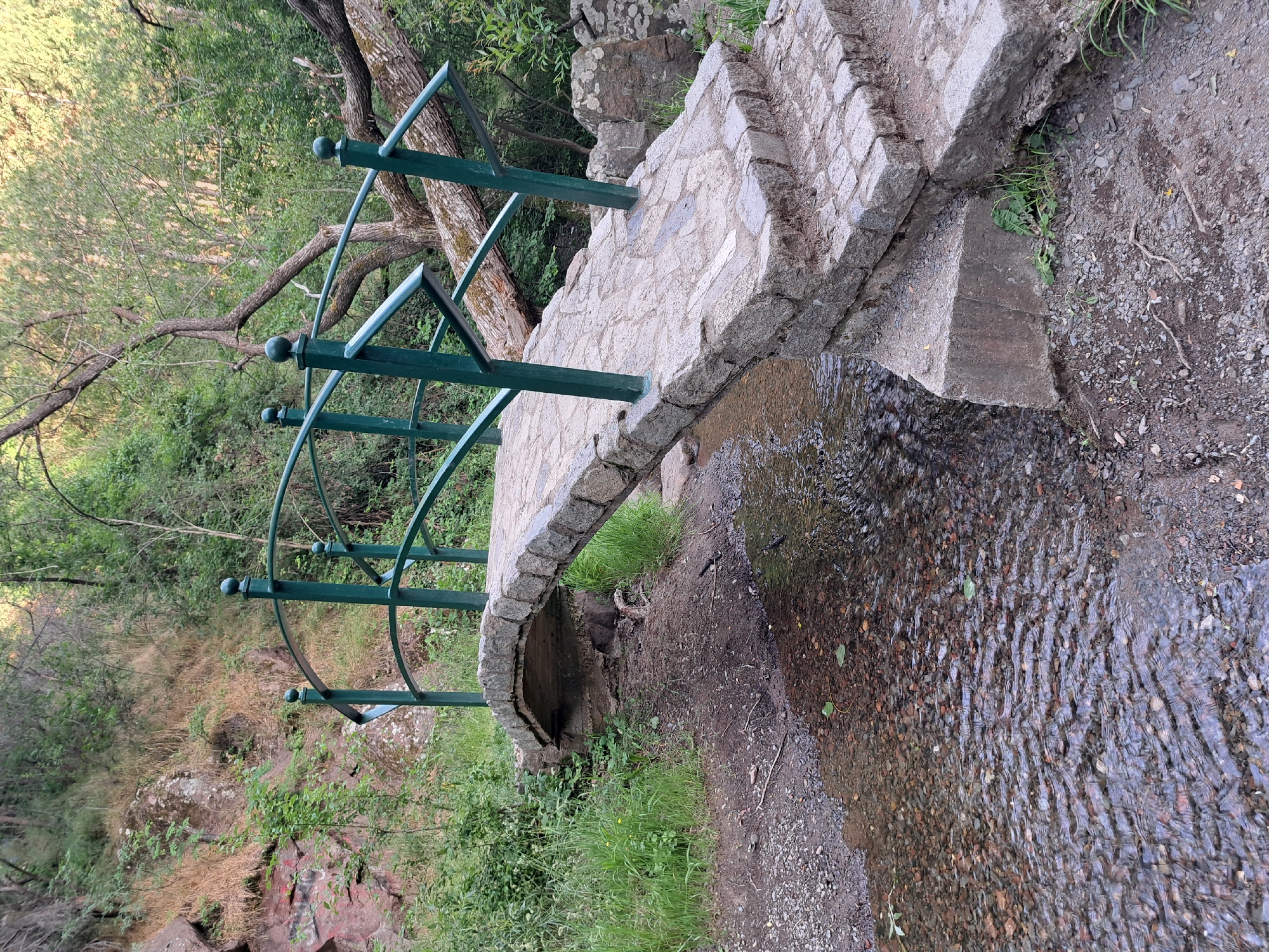 A stone bridge spans a shallow, clear stream flowing over a rocky bed in a wooded area. The bridge is constructed with rough-hewn stones and features a green metal railing. Steps made of similar stone lead up to the bridge on the right side. The surrounding environment is lush with green vegetation, including trees and grass, and a rocky hillside is visible in the background. The scene appears to be a natural setting within or near the city of Lokorsko, Bulgaria.