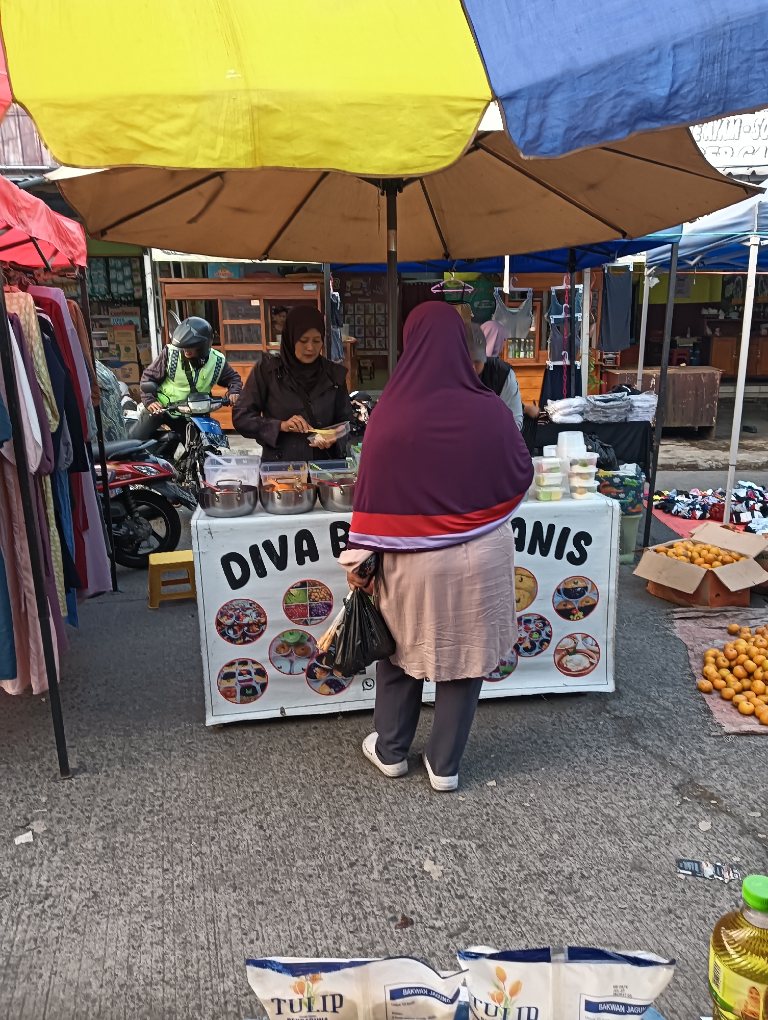 In Katapang, Indonesia, an outdoor market scene unfolds under a yellow and blue umbrella. A woman wearing a purple hijab and a light-colored skirt stands with her back to the camera, holding a plastic bag. She is facing a food stall with a white board displaying the words "DIVA BANANIS" and circular images of various food items. Behind the stall, a woman in a brown hijab is serving food, and a man wearing a neon green vest and helmet is on a motorcycle. The ground is paved with asphalt, and several boxes of oranges are visible to the right, with more scattered on the ground. Clothing racks with garments are set up to the left of the stall. In the foreground, two bags of what appears to be flour are placed on the ground.
