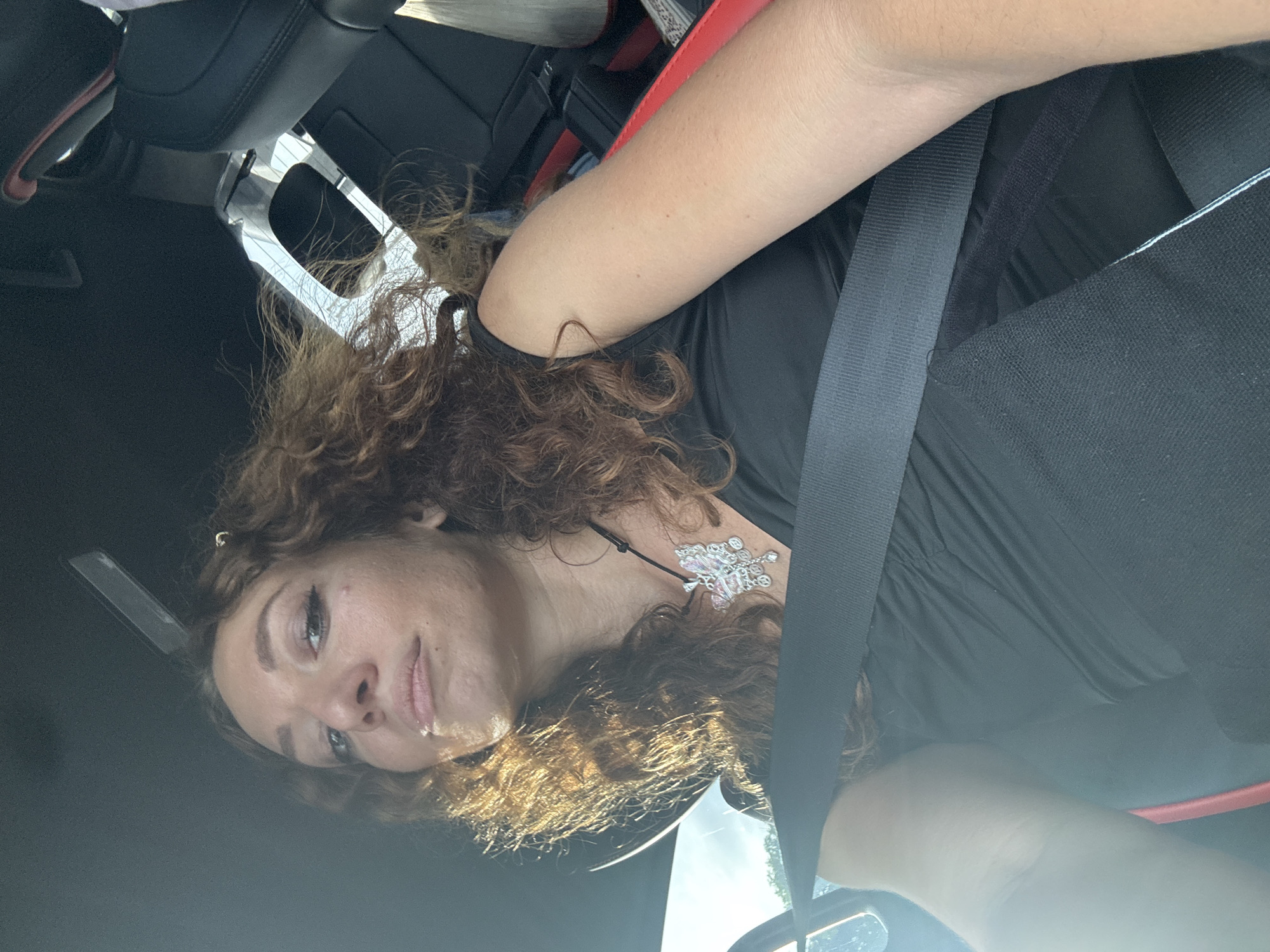 A medium shot captures a woman with curly brown hair, wearing a black top and a silver butterfly pendant, seated in the passenger seat of a car. She is looking upwards and slightly to the left, her mouth slightly open. A seatbelt is diagonally across her chest. The interior of the car is visible, including the dashboard and a portion of the passenger seat. The scene appears to be lit by natural light coming from outside the car, casting shadows and highlights on the woman's face and hair. The location is not specified in the image or provided context.