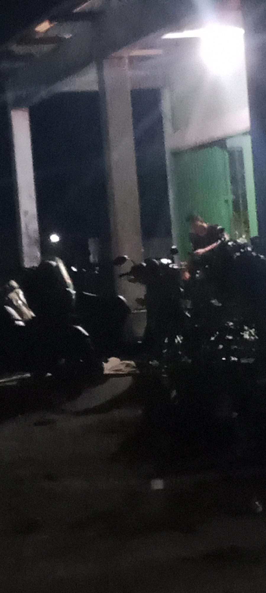 The image shows a dark outdoor scene, likely at night, with a person standing in front of a green door. Several motorcycles are parked in the foreground. The location is described as Kerumutan, Indonesia, though specific landmarks are not visible. The lighting is dim, with a bright light source in the background illuminating the scene. The person in the image appears to be looking away from the camera. The overall impression is of a casual, everyday scene.