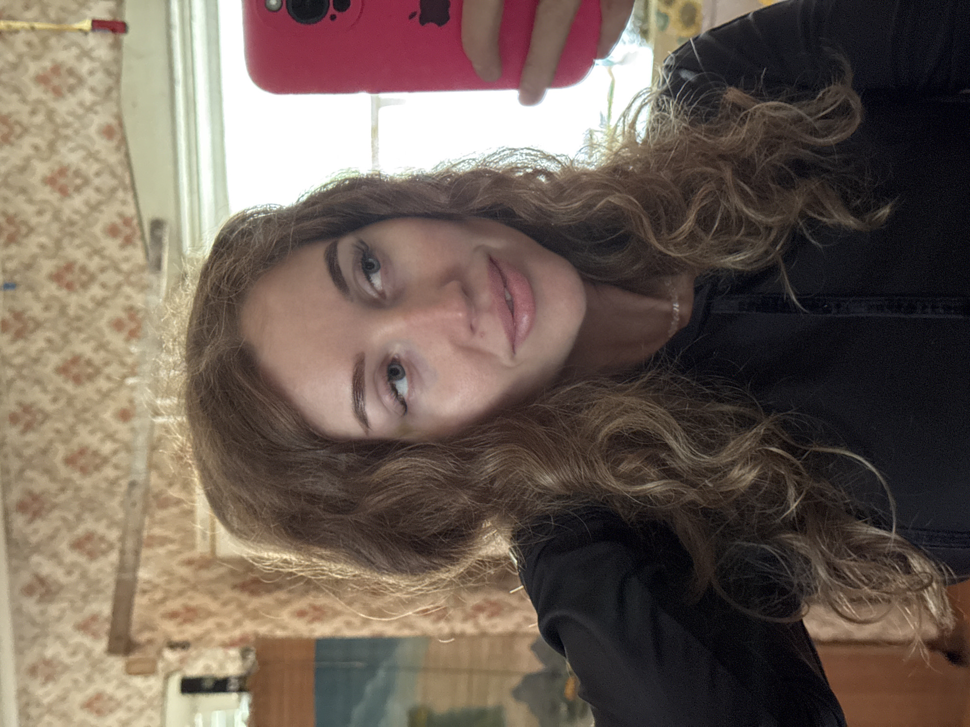 A selfie-style portrait features a young woman with long, curly light brown hair, appearing to hold a pink smartphone with an Apple logo above her head to capture the image. She has light-colored eyes and is looking directly at the camera with a neutral to slight smile. She wears a dark, possibly black, long-sleeved top and a silver ring on her left hand.
The background reveals an indoor setting, likely a room. Prominently featured is light-colored wallpaper with a recurring floral or geometric pattern in shades of beige and reddish-brown. A bright window is visible in the upper background, indicating daytime and providing natural light. Below the window, a portion of what appears to be a framed landscape painting or picture, featuring blue and green tones, can be seen. Another faint yellow, possibly floral, pattern is visible near the top right of the frame, behind the phone. The overall ambiance suggests a domestic interior in Ledukhi, Russia.