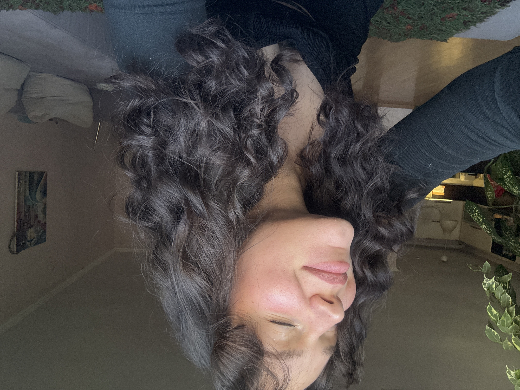 A woman with long, dark, curly hair is the focal point. She is wearing a black long-sleeved top and black pants. Her eyes are closed. Behind her are interior elements, including a sofa, a wall with a picture, a countertop, and plants. The image doesn't contain information to identify the location as Burgas, Bulgaria.