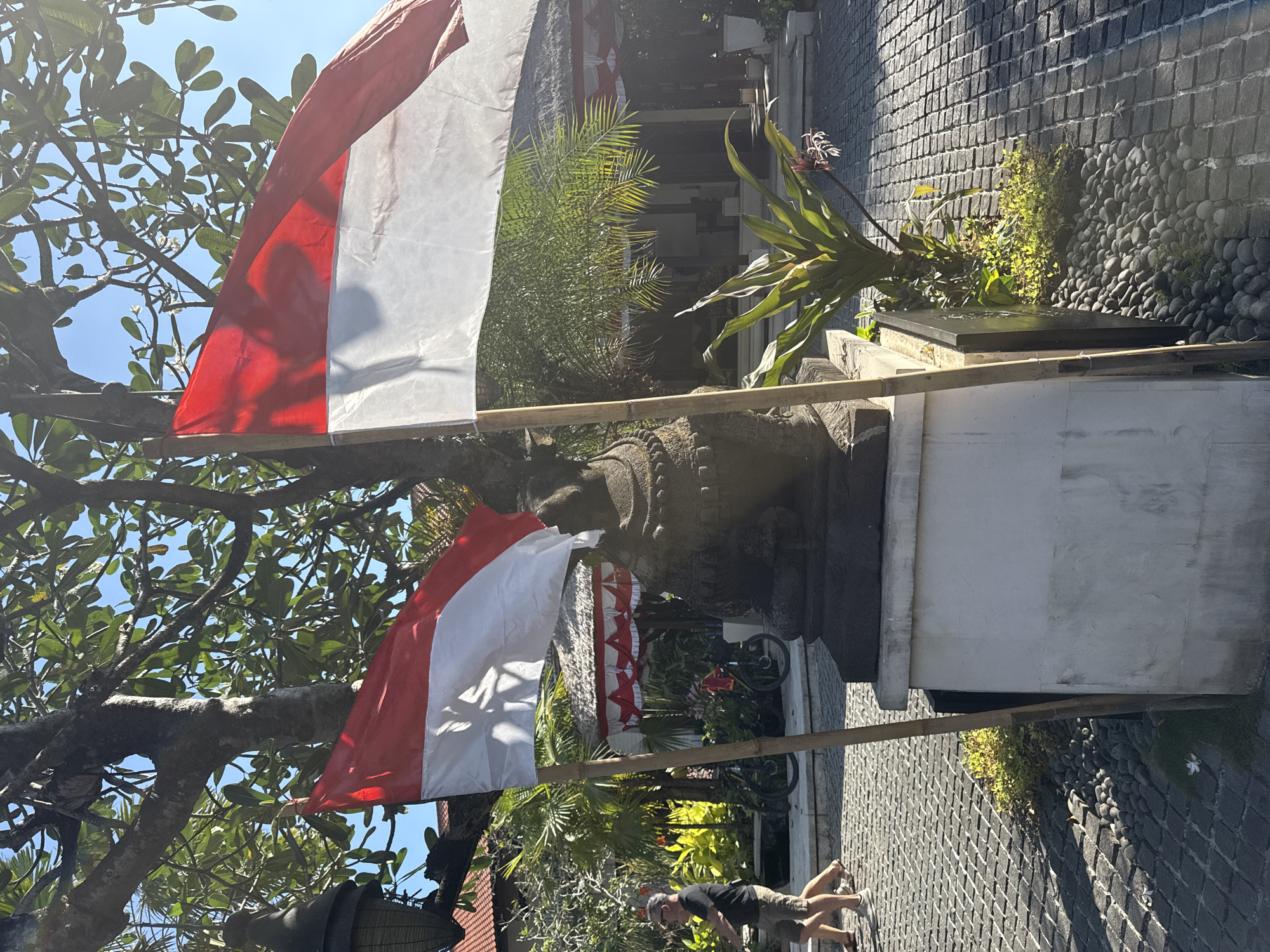 Two Indonesian flags, red over white, are prominently displayed on bamboo poles in the foreground. A stone statue, likely a cultural representation, sits on a pedestal between the flags. Behind the flags and statue, a paved walkway leads to a building with a thatched roof and red and white decorative awnings. Green foliage, including palm fronds and other plants, surrounds the area. Two individuals are walking on the paved path in the middle ground, and bicycles are visible in the background. The scene is set outdoors with a clear blue sky overhead, suggesting it is a sunny day. The architecture and flags indicate the location is likely in Denpasar, Indonesia, potentially at a hotel, cultural site, or public building.