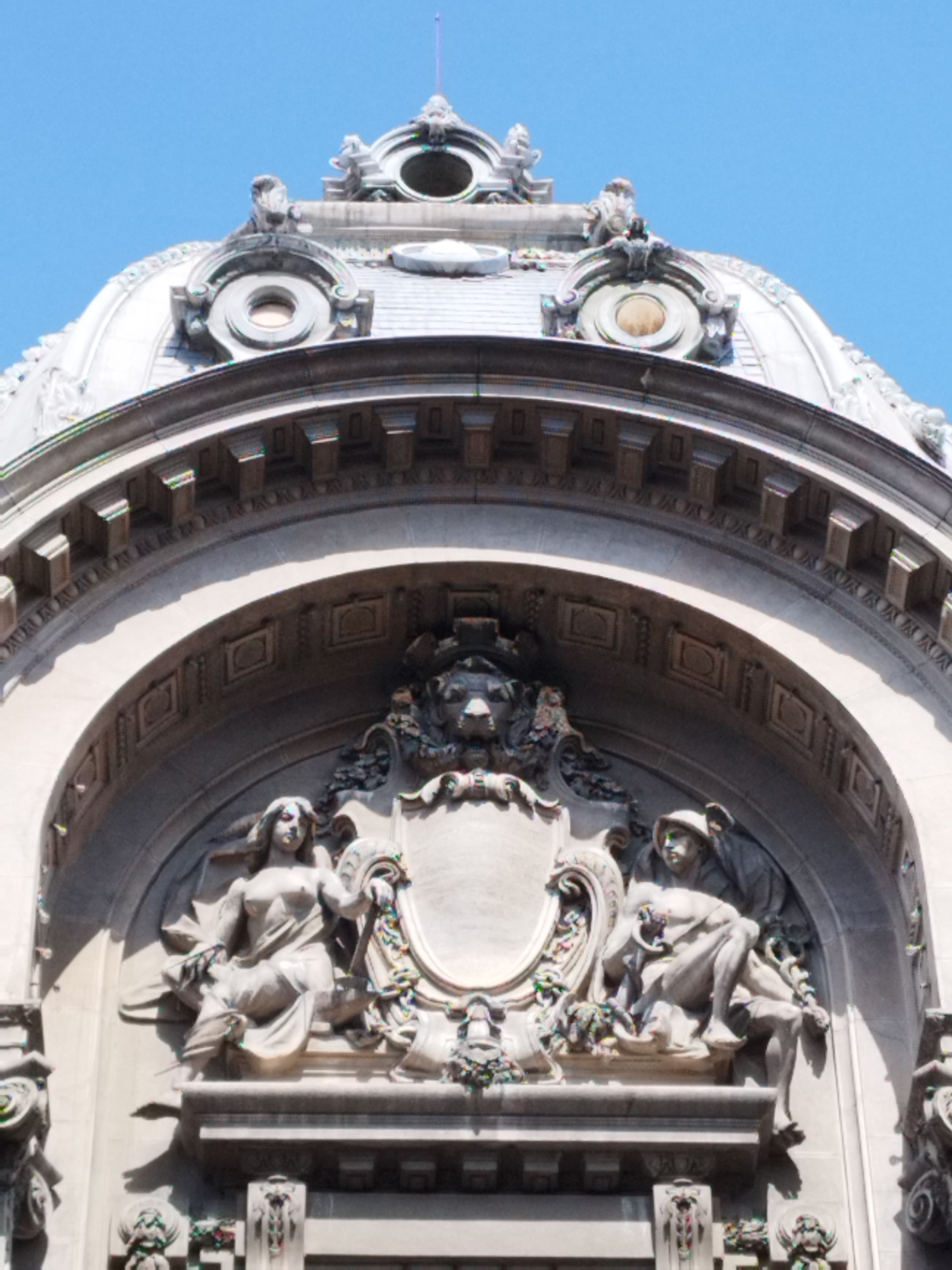 This is a detailed, eye-level, medium shot of the facade of the CEC Palace in Bucharest, Romania. The image captures an ornate architectural element featuring two sculptures above a large archway. The sky above is a clear, bright blue, suggesting it is daytime with good weather.
The central focus is a semi-circular tympanum filled with intricate bas-relief sculptures. At the apex of this tympanum is a lion's head carved in stone, appearing fierce with its mouth open. Flanking this lion's head are decorative floral elements and scrolls. Below the lion's head, a blank stone cartouche or escutcheon is positioned.
On either side of this central decoration, within the archway, are two larger, full-body sculptures. The sculpture on the left depicts a female figure seated with her legs crossed. She is draped in flowing robes, with her upper body exposed. She holds a palette and brush in her left hand and rests her right arm on a shield or crest. The sculpture on the right shows a male figure, also seated, wearing a helmet and a cloak. He holds a snake coiled around his arm. Both figures appear to be seated on decorative pedestals adorned with more floral carvings and scrolls.
The overall architectural style is grand and ornate, characteristic of Beaux-Arts architecture, with extensive use of stone carvings, moldings, and classical motifs. The material appears to be light-colored stone, possibly limestone or marble, which has weathered over time. The lighting suggests the sun is casting shadows, particularly on the right side of the image, highlighting the three-dimensional nature of the sculptures and architectural details. The photograph appears to be taken from a slightly upward angle, looking towards the upper part of the building's facade.