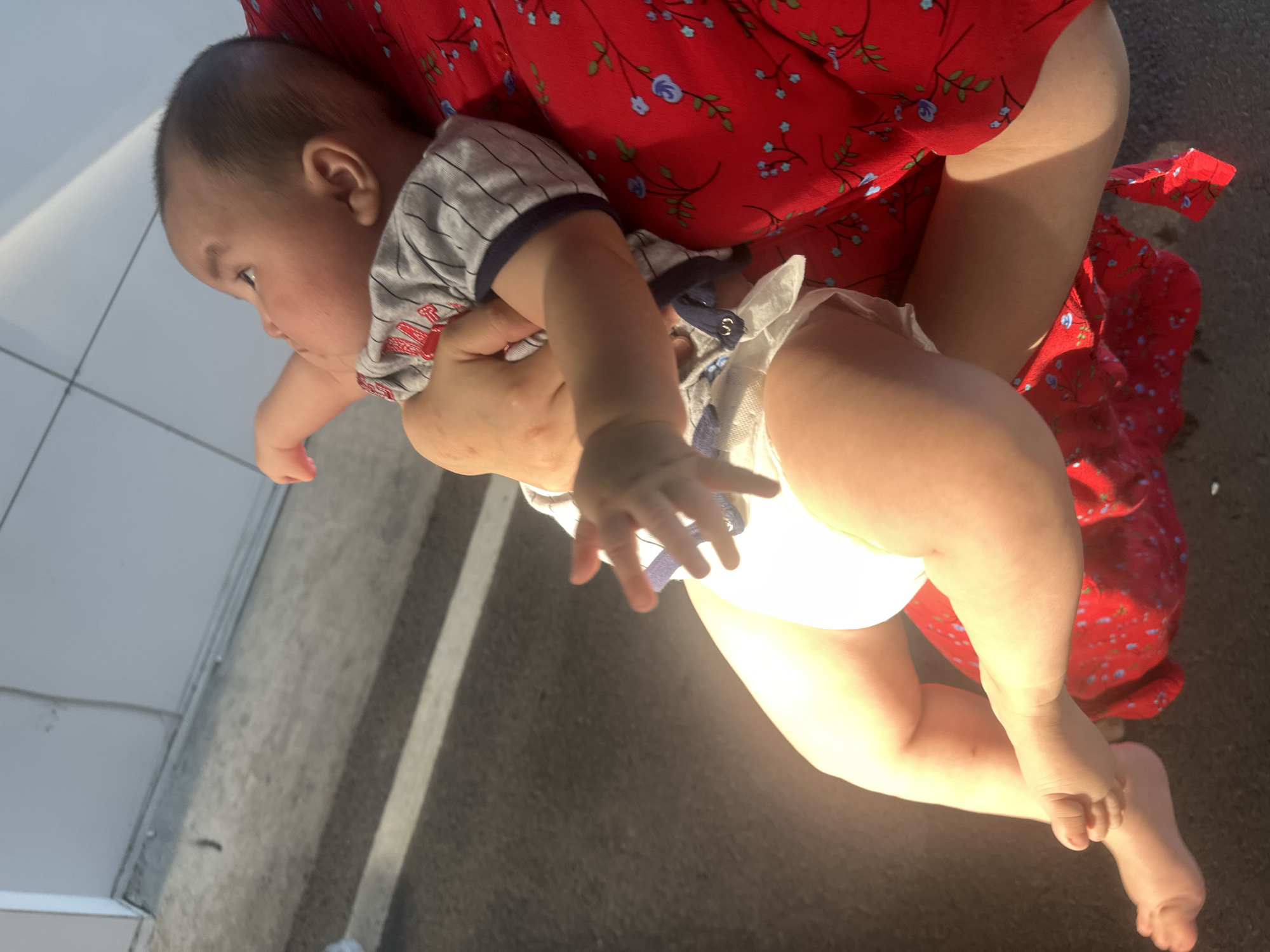 In this image, a baby is being held by an adult, likely a parent or caregiver, dressed in a red floral print dress. The baby is wearing a grey and white striped t-shirt and a diaper. The scene appears to be outdoors, with a grey, textured surface that could be asphalt or concrete in the foreground, marked by two white parallel lines. To the left, a light grey tiled wall or surface is visible, possibly part of a building or walkway. The lighting suggests it is daytime, with a strong light source casting shadows and highlighting the baby's legs. The overall impression is a casual, everyday moment. The setting is identified as Manas, Kyrgyzstan. There is no visible text in the image.