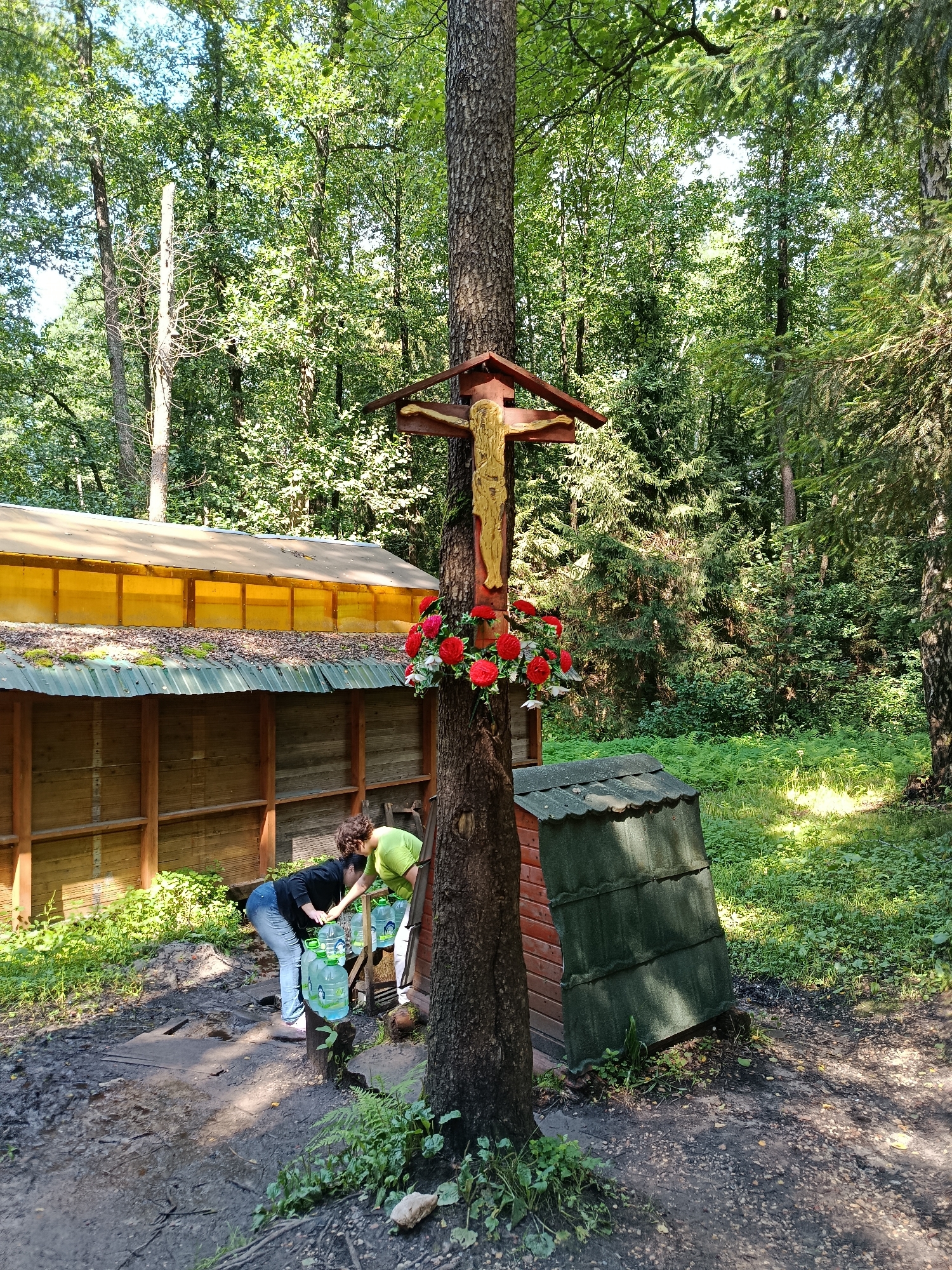 In Staryi Pokrov, Russia, within a wooded area, a Christian cross with a depiction of Jesus is mounted on a tree, adorned with red artificial flowers. Two women are present near a wooden structure with a yellow translucent roof and a green tiled roof. One woman, wearing a black jacket and blue jeans, is bent over, reaching for a large plastic water bottle. The other woman, in a light green shirt, is also leaning down, assisting with the bottles, which are arranged on a wooden stand. The ground is muddy and uneven, with scattered leaves and vegetation. The surrounding environment is dense with trees and foliage, suggesting a natural or semi-natural setting.