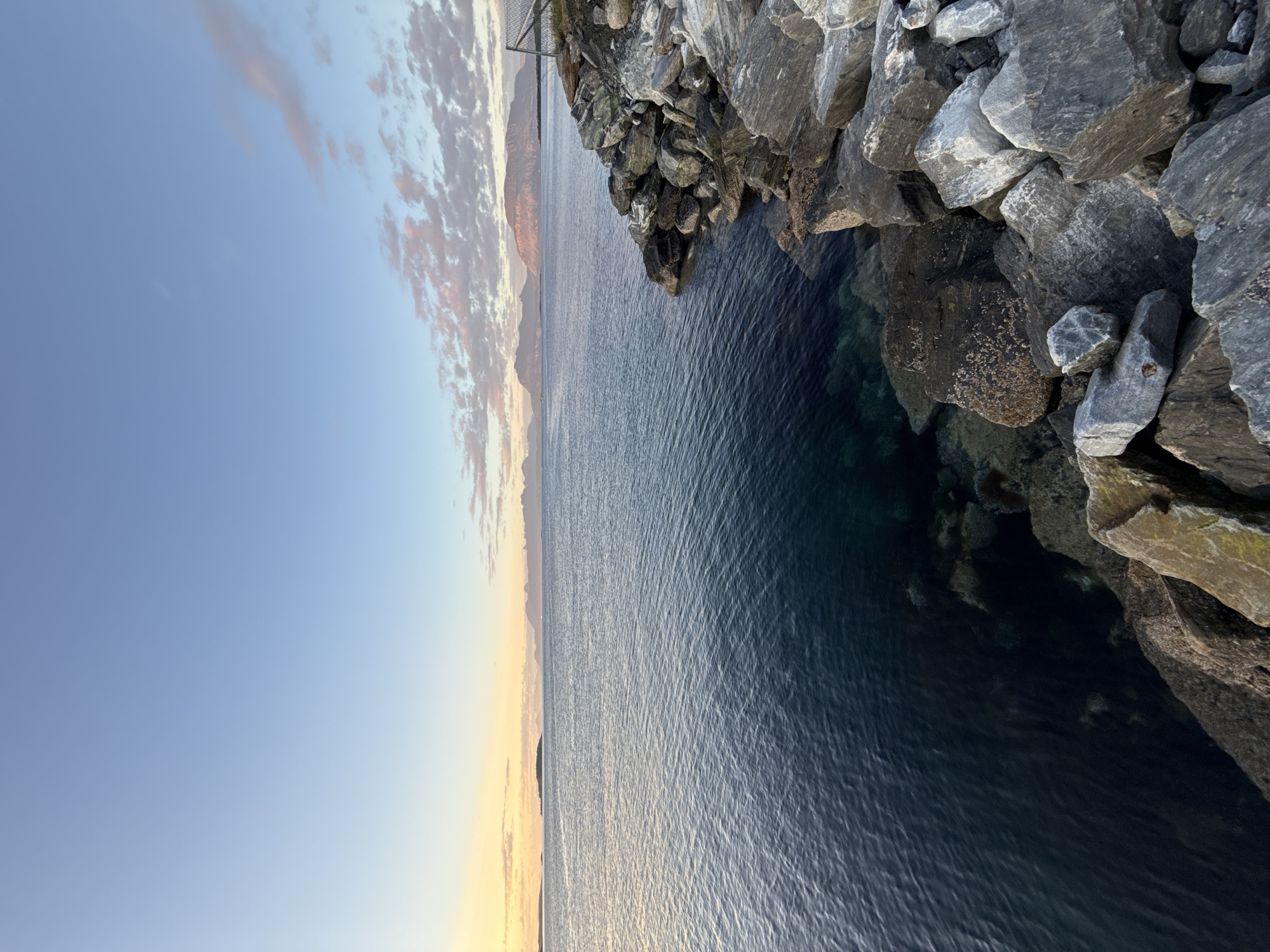 A wide shot captures a serene seascape at dusk, with the sun setting behind distant mountains. The foreground is dominated by a rocky shoreline, with large grey and tan boulders piled against the dark, rippling water. The water itself is a deep blue, with hints of green visible near the shore, suggesting shallowness and clarity.
In the middle ground, the vast expanse of the sea stretches towards the horizon, where a faint line of islands and the silhouette of more distant, hazy mountains are visible. The sky above is a gradient of pale blue, transitioning to soft pinks and yellows near the horizon, with wispy clouds catching the last light of the day. A simple wire fence is visible on the right side of the frame, marking the edge of the land.
The scene evokes a sense of tranquility and the natural beauty of the Norwegian coast. The lighting suggests that this image was captured during the golden hour, just before twilight. Based on the presence of the characteristic mountain ranges and the general coastal landscape, this scene is consistent with the area around Harstad, Norway, likely overlooking the Norwegian Sea. There are no people or active elements present in the image, focusing solely on the natural environment.