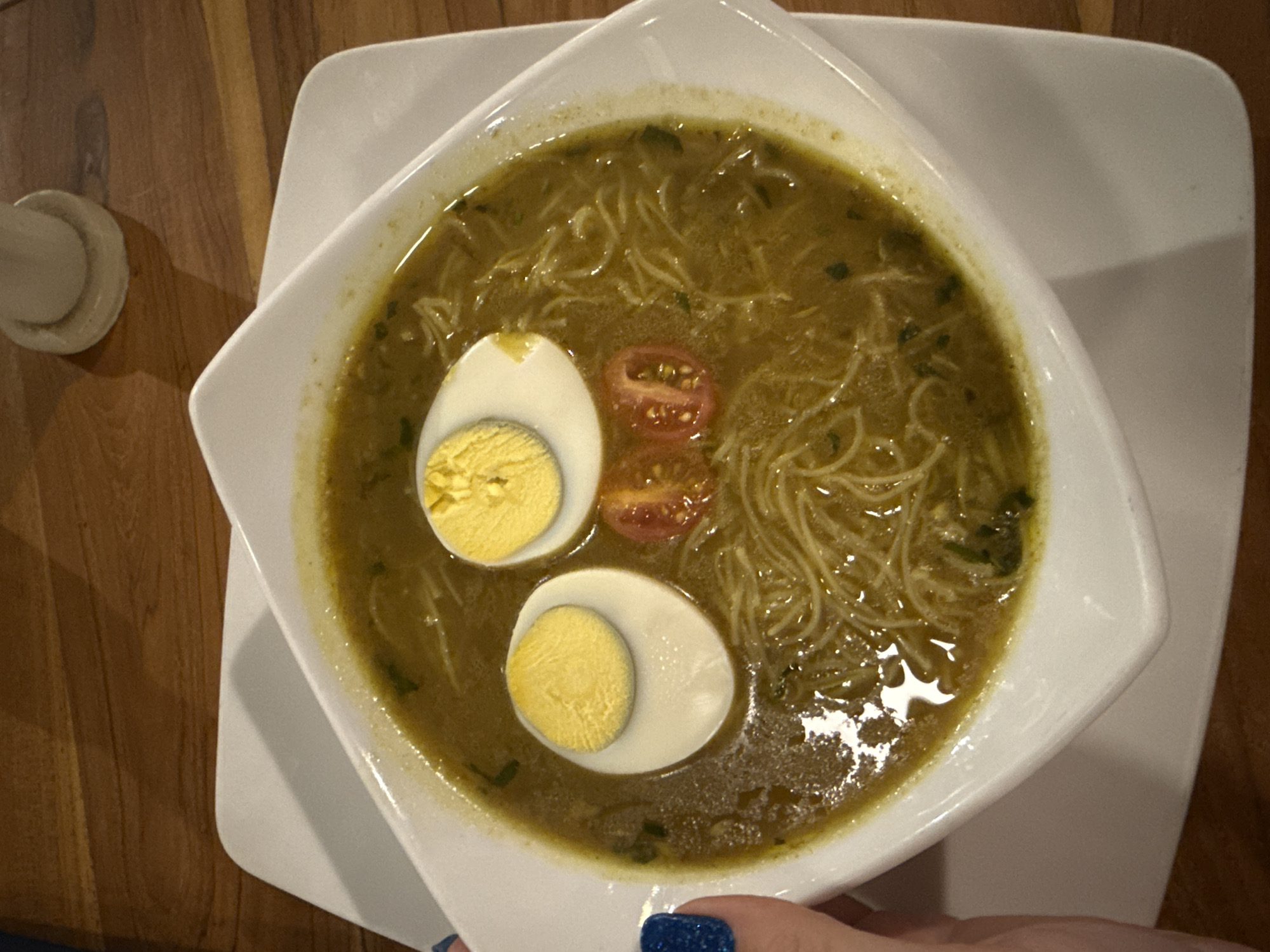 A close-up, top-down shot shows a white square bowl filled with a greenish-yellow broth containing thin noodles. Two halves of a hard-boiled egg are placed in the soup as eyes, and two halves of cherry tomatoes are positioned below them as a mouth, creating a smiley face. A hand with blue, glittery nail polish is visible on the left side of the frame, reaching for the bowl. The bowl sits on a larger white plate, which is placed on a wooden surface. A small white cylindrical object is also visible on the wooden surface in the upper right corner. The scene appears to be indoors, likely in a restaurant or dining setting within Denpasar, Indonesia, given the context of the request.