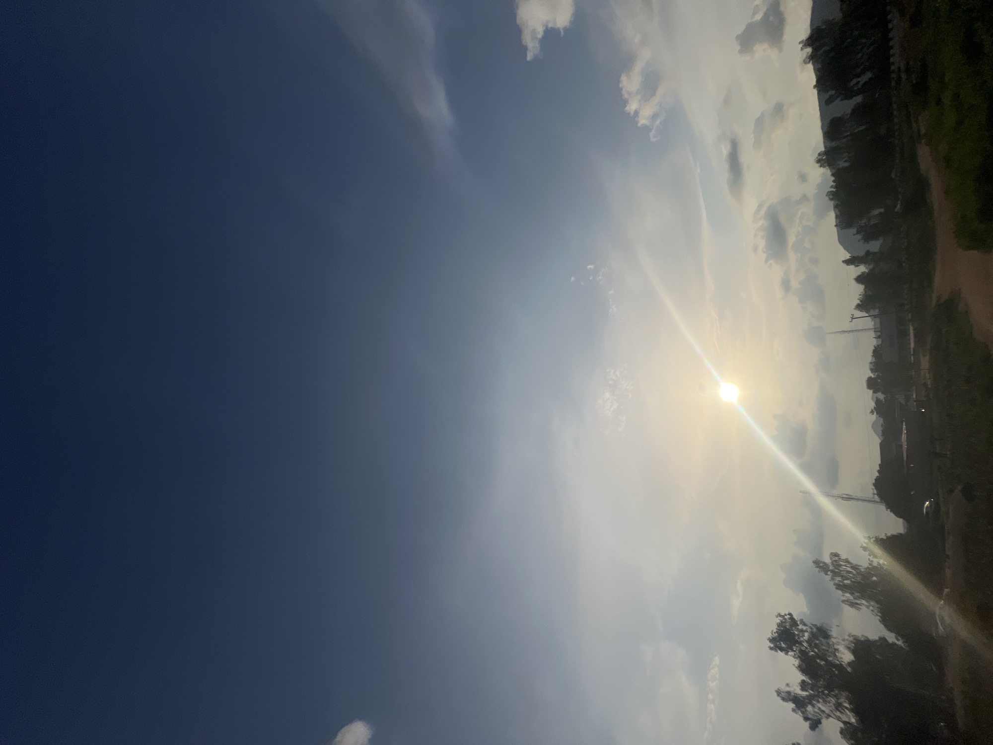 The image depicts a sunlit sky over a rural landscape, likely in Howayi, Nigeria. The sun is prominently positioned in the upper-middle section of the frame, casting a bright glare and a visible light ray across the scene. The sky transitions from a deep blue at the top to lighter shades of blue and white, with scattered clouds present.
In the lower portion of the image, a treeline of silhouetted trees marks the horizon. Beyond the trees, there are hints of structures, suggesting a small settlement or scattered buildings. A dirt path is visible in the foreground, winding through grassy terrain. A few vehicles are partially visible amongst the trees, and a tall tower, possibly a utility pole or communication tower, stands out against the sky. The overall impression is that of a serene, natural environment under a bright sky, possibly during late afternoon or early morning given the quality of light.