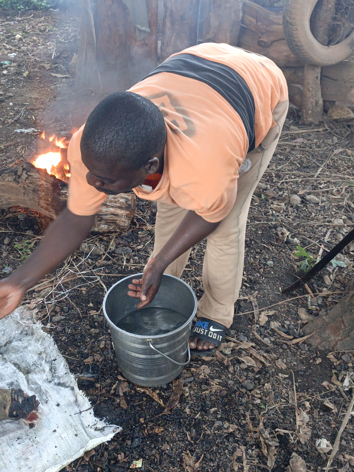 A man stands outdoors in Akwanga, Nigeria, next to a fire, bending over a metal bucket filled with water. He is wearing an orange T-shirt with a black band, beige pants, and black Nike sandals. His hands are near the bucket, possibly washing them. Beside the bucket is a stained, white cloth. The ground is covered in loose soil, leaves, and small rocks. A stack of wood and a tire hang from wooden posts in the background.