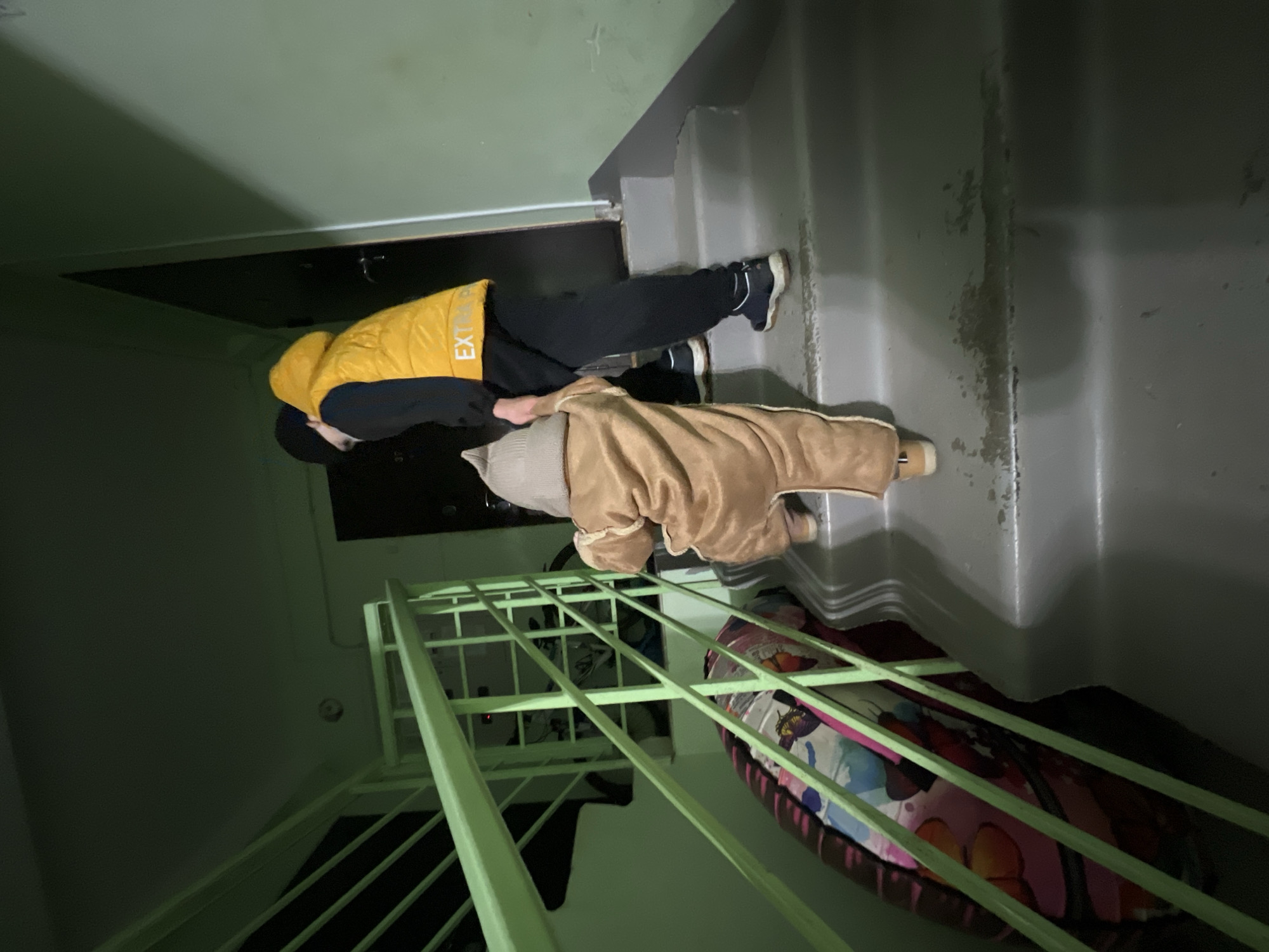 A person wearing a yellow vest with "EXT" visible on the back, black pants, and dark shoes is lying on a gray staircase. The person is possibly holding or lying on top of a person wearing a tan outfit and light-colored shoes. The scene appears to be indoors, possibly in a stairwell with light-green painted railings. A colorful object sits below the railing, possibly a folded comforter. The exact location is unknown without further details, as "Noviy, Russia" is too broad.