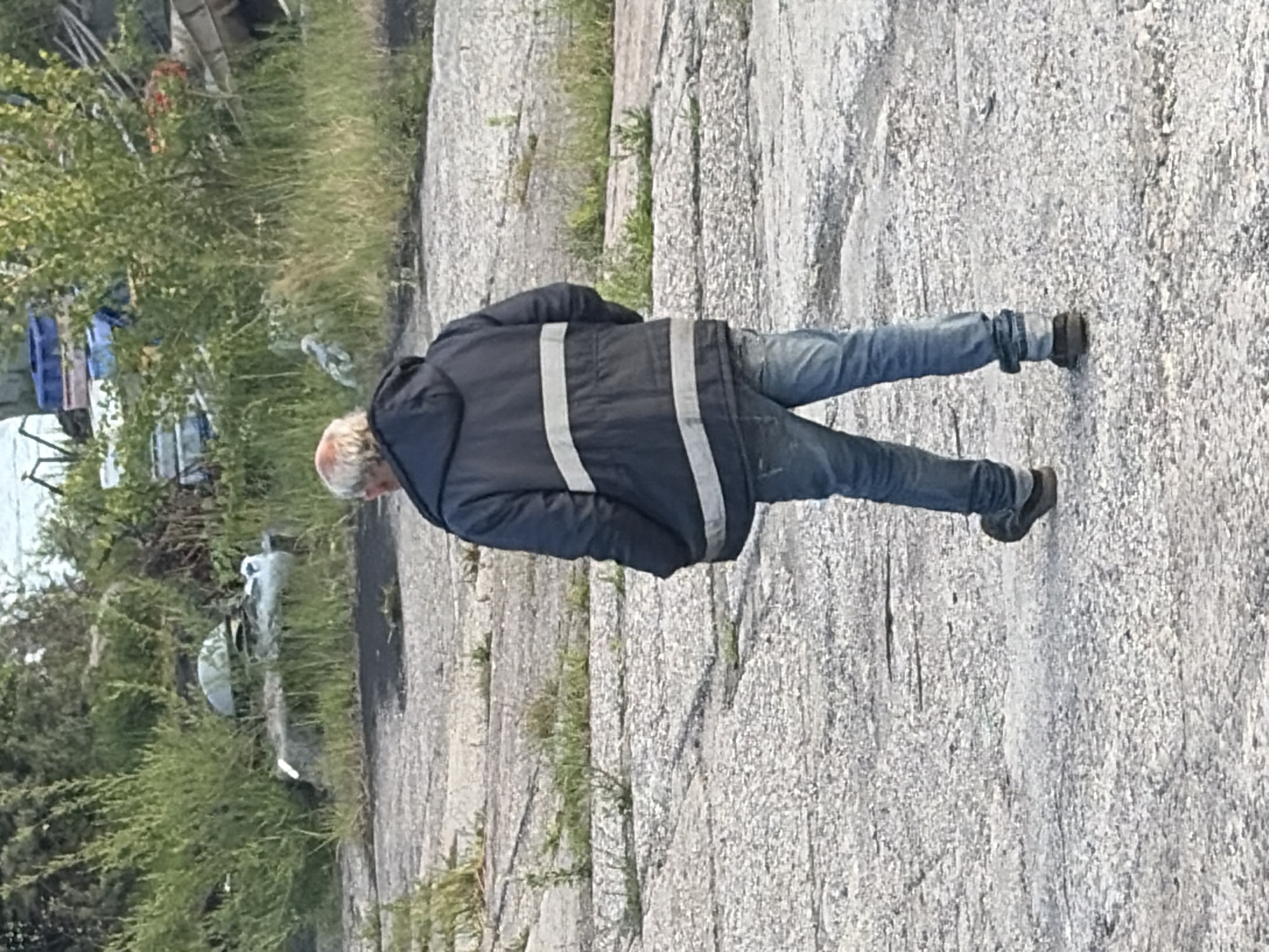 A middle-aged man with graying hair is walking away from the camera on a cracked concrete surface. He is wearing a dark blue hooded jacket with two horizontal white reflective stripes across the chest and sleeves, and blue jeans. The background consists of overgrown green grass, bushes, and debris, including some blue plastic containers and white objects. The scene appears to be an outdoor, possibly industrial or undeveloped area, with a body of water visible in the distance. The location is specified as Baltiysk, Russia.