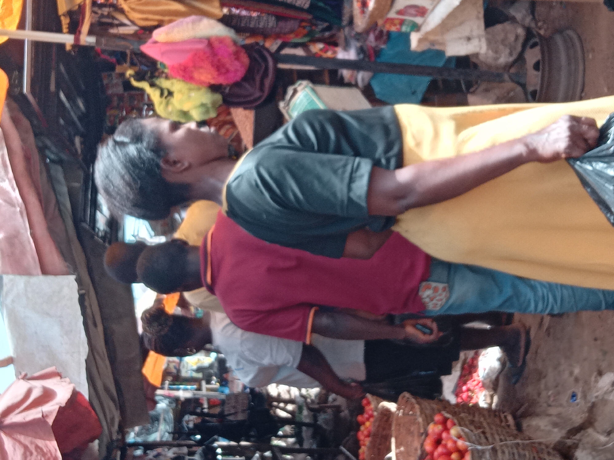 A street-level view of a crowded market in Sabon Tasha, Nigeria. People are shopping; the woman nearest the camera wears a black short-sleeved shirt over a yellow dress, holding a black plastic bag. Behind her, a man is dressed in a maroon shirt, followed by someone in a white shirt. Wicker baskets containing red tomatoes are prominent in the foreground. Textiles and merchandise hang overhead.