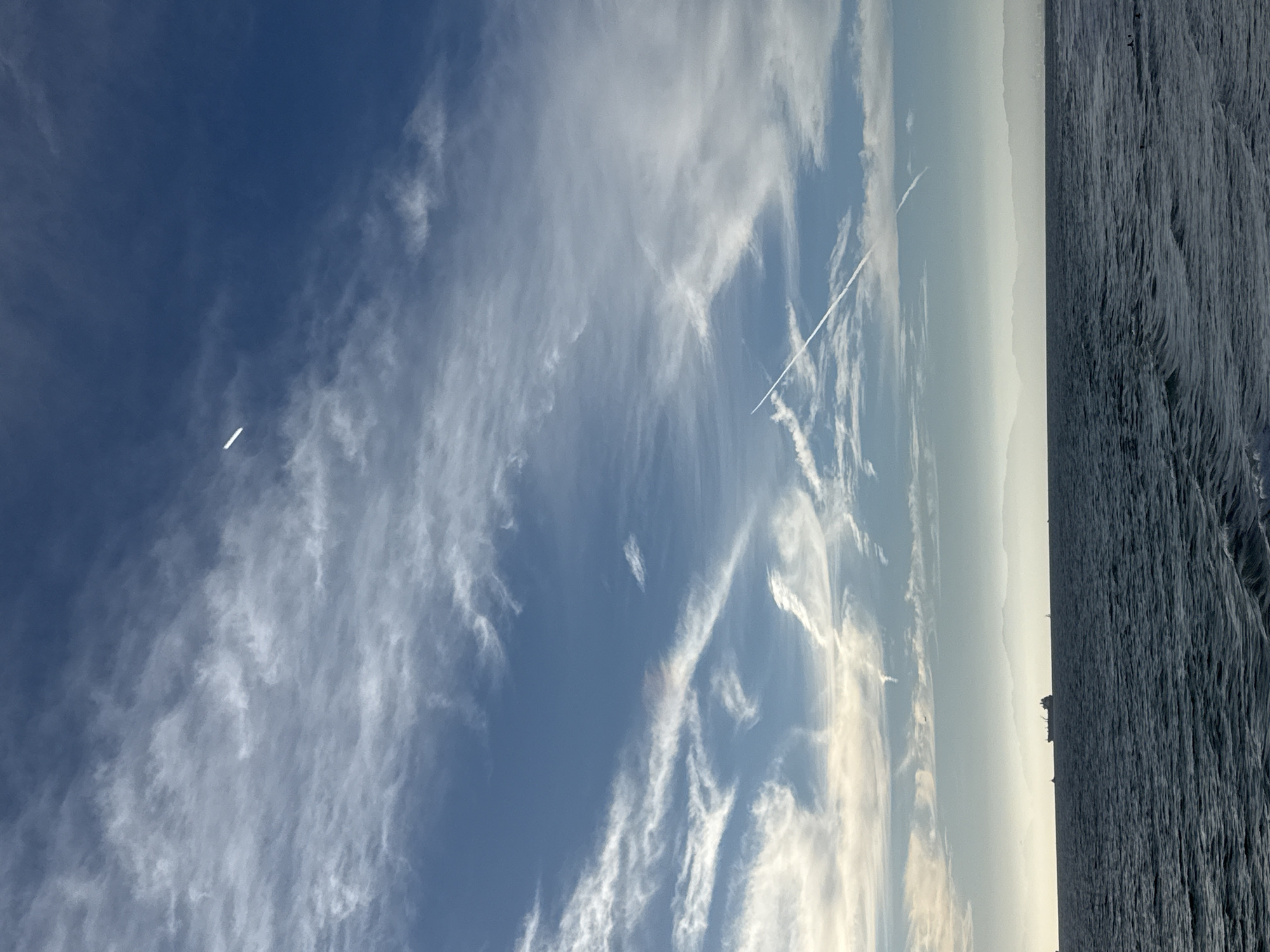 This image depicts a view of the ocean and sky from Manhattan Beach, California. The scene is dominated by a vast blue sky filled with wispy white clouds. There are two visible contrails from airplanes in the sky, one long and diagonal and another shorter one. In the distance, a hazy mountain range is visible on the horizon. In the foreground, the dark blue ocean has small waves, and a large ship is sailing on the water. Several smaller boats are also visible on the water. The overall impression is one of a serene and expansive coastal landscape.