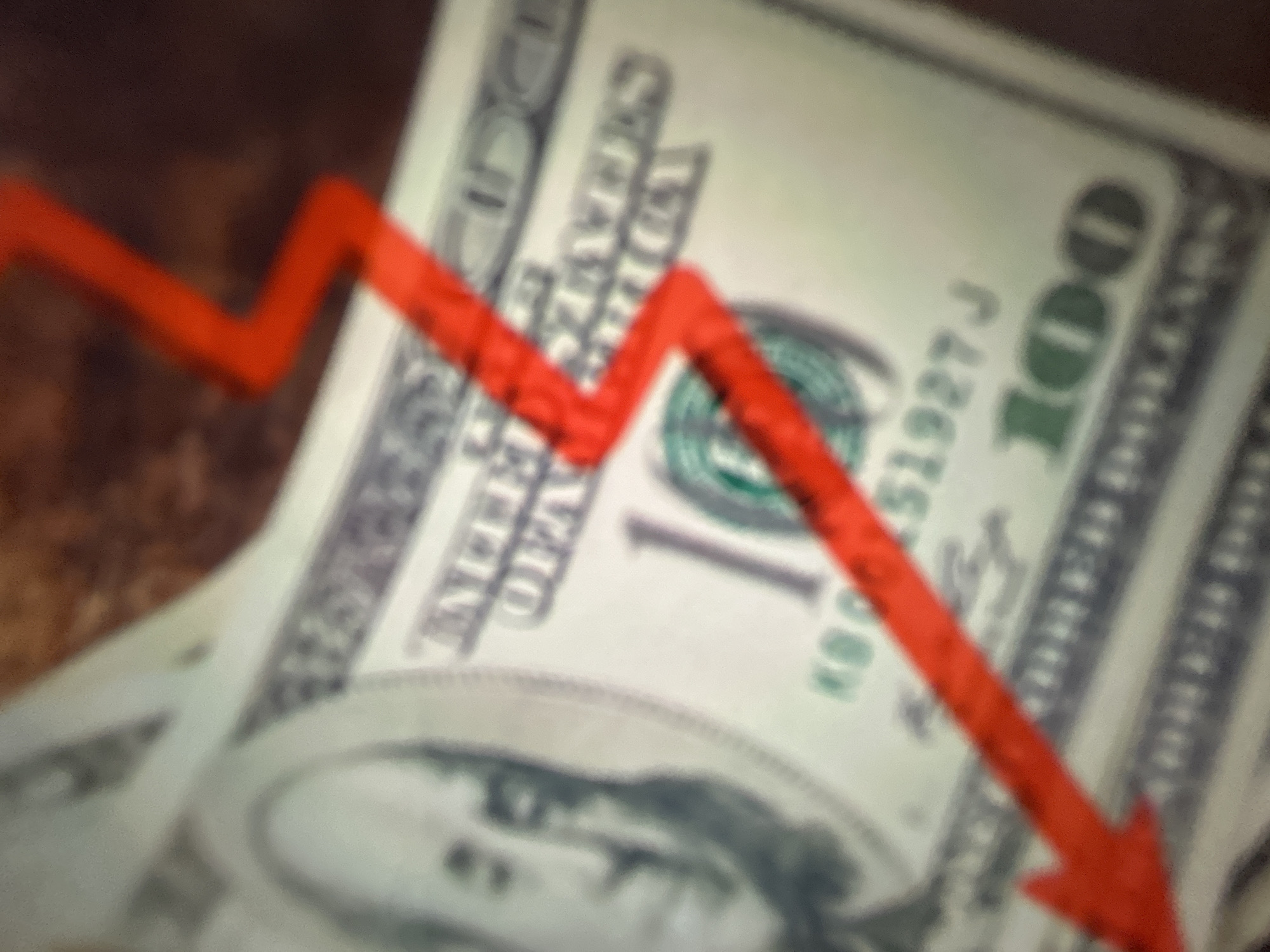 The image shows a close-up, out-of-focus view of US one hundred dollar bills. A thick red arrow, creating a zig-zag pattern, overlays the money, pointing downwards. The arrow appears to indicate a financial decline or loss. The bills themselves are partially obscured by the arrow, but the "100" denomination and parts of the portrait of Benjamin Franklin are discernible. The background is a blurred, dark surface. No specific time of day, weather, or strong emotional cues are evident. No visible text related to Gelendzhik, Russia is present.