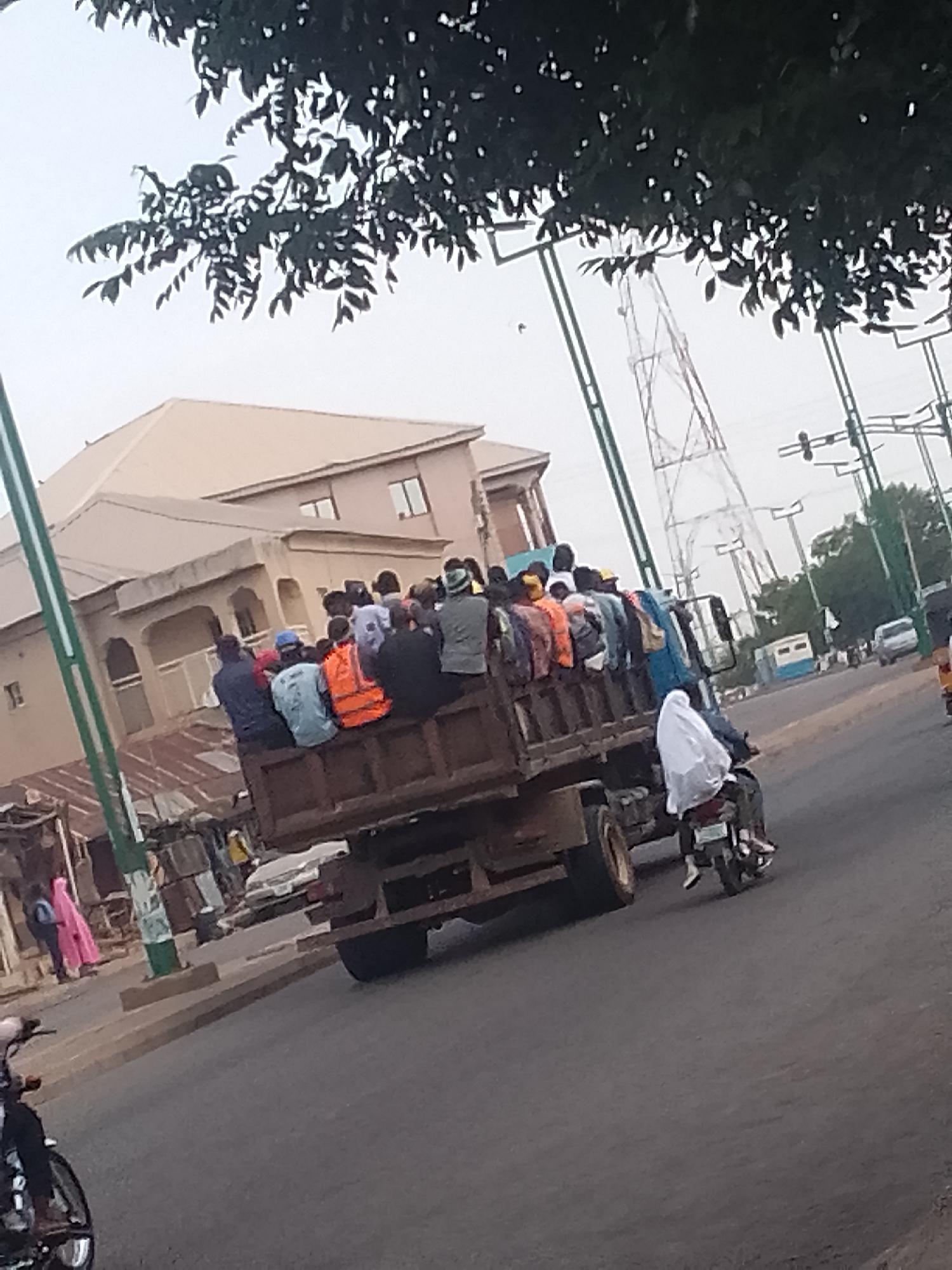 In Arawa, Nigeria, a large truck is overloaded with over two dozen people, many of whom are sitting on the sides of the truck bed. The truck is traveling on a paved road, with a building and trees visible in the background. A person on a motorcycle, wearing a white veil, is riding next to the truck. The overall scene depicts a common form of transportation in the region, highlighting overcrowding and potential safety concerns.