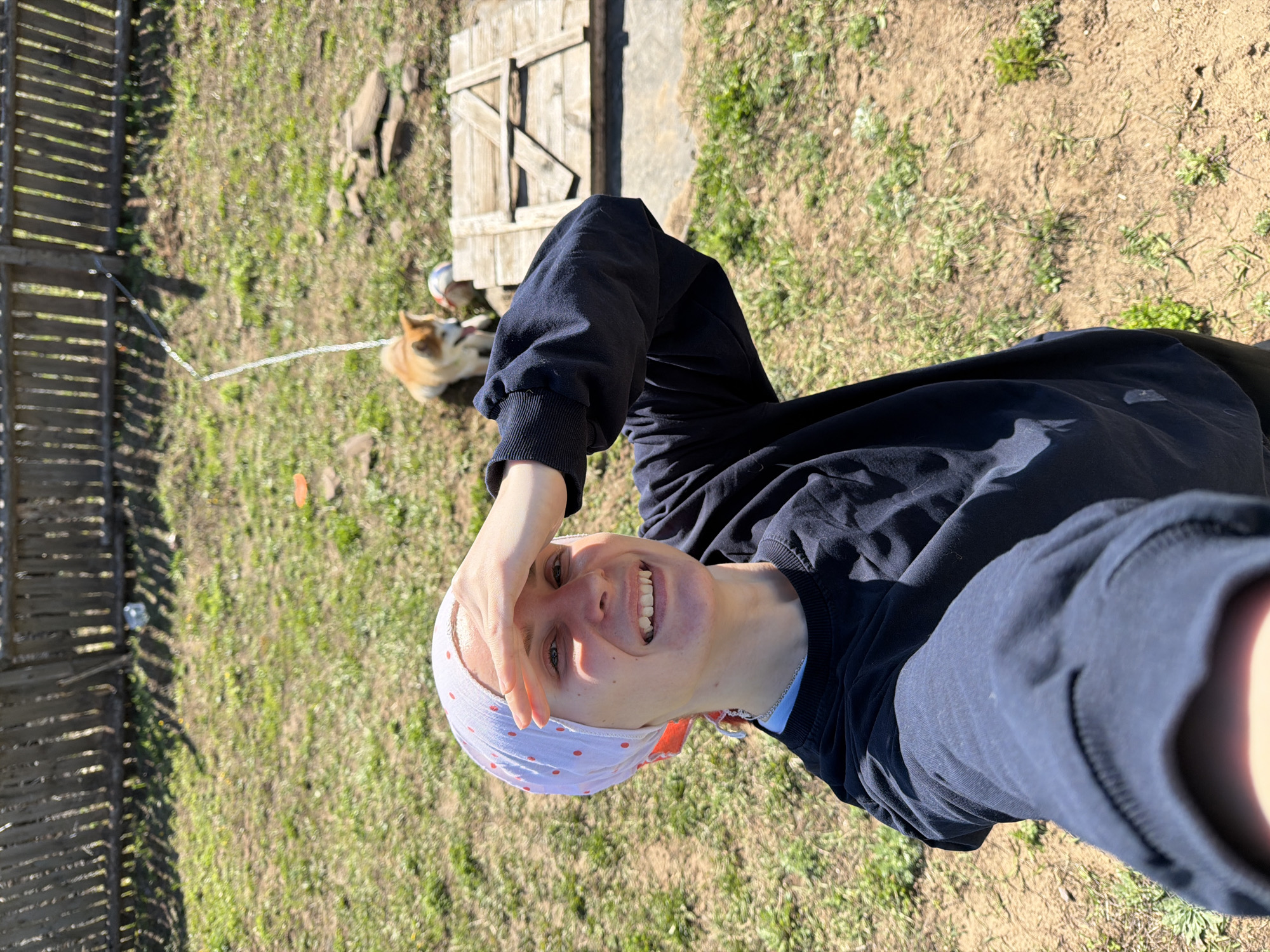 A person, smiling with their hand over their head, is captured in an outdoor setting in what might be Chaikovsky, Russia. The person is wearing a dark sweatshirt and a white headscarf with red polka dots. A dog on a leash is visible in the background, along with a wooden fence, wooden structure, and grass. The scene is illuminated by sunlight.