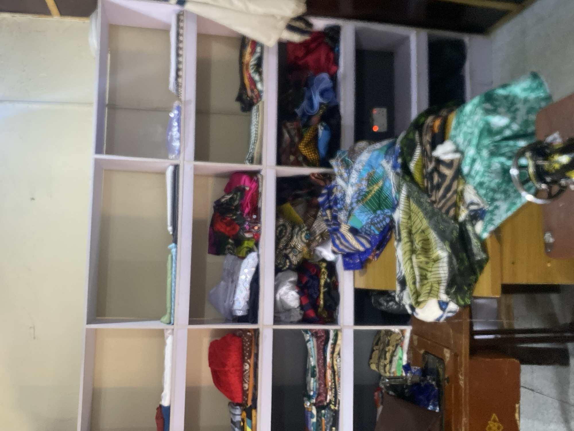 The image shows a room in Bukuru, Nigeria, with shelves filled with folded fabrics, primarily in vibrant colors and patterns. The fabrics appear to be of various types, suggesting a textile or tailoring business. A sewing machine is visible on a wooden table in the foreground, along with more stacks of fabric. The shelves are light-colored, and the walls are a pale yellow. The overall impression is of an organized storage space for materials used in sewing or fashion design. The lighting is subdued, creating a somewhat dim atmosphere.