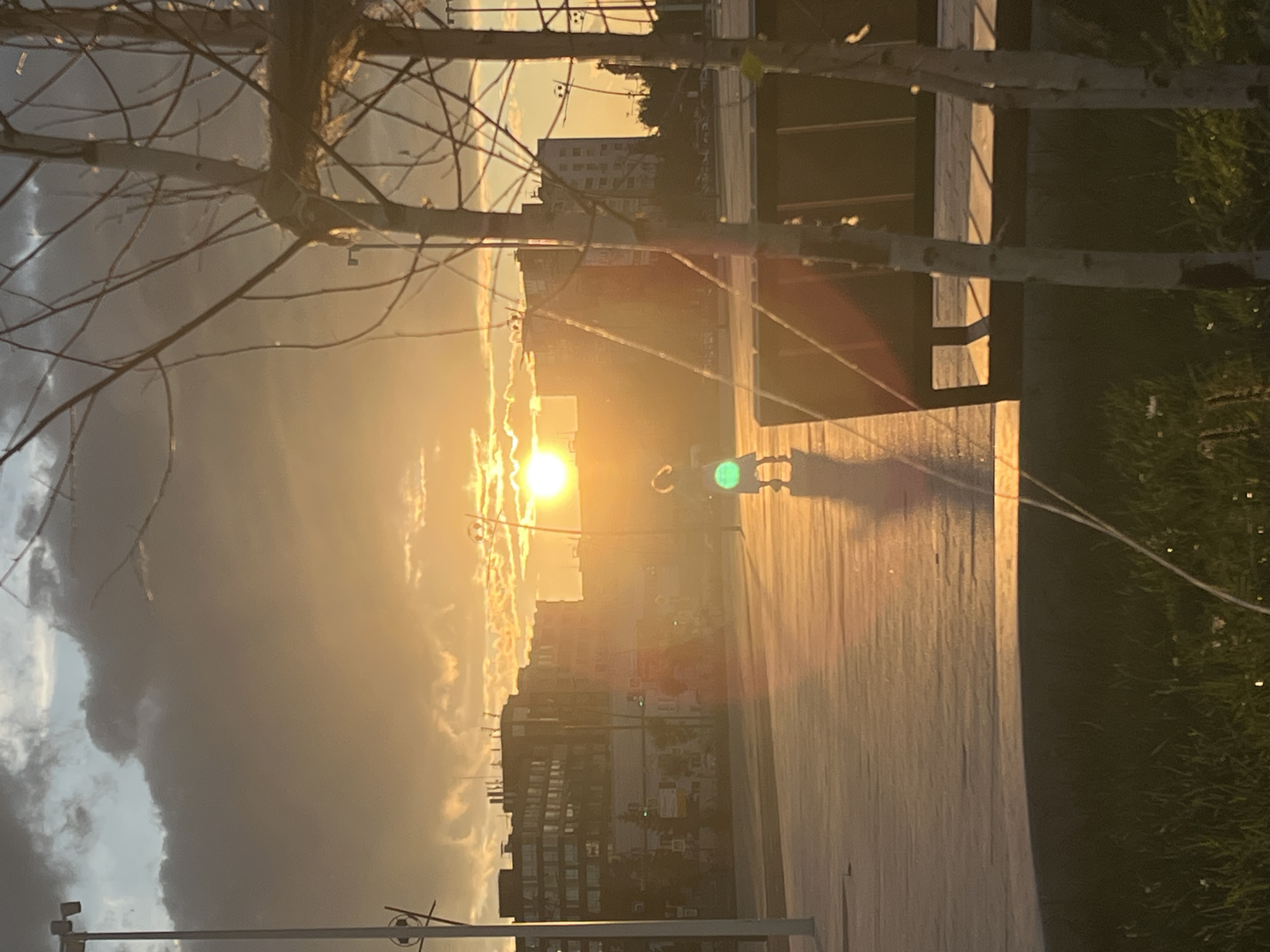 The image depicts a sunset over a modern urban environment in Nizhny Novgorod, Russia. The sun is low on the horizon, casting a warm, golden glow across the scene and creating long shadows. In the foreground, two young trees with bare branches are silhouetted against the light. To the right, a dark park bench is visible. The middle ground features a paved open area where a child, appearing to be in motion, is illuminated by the setting sun. The child is wearing dark clothing and a bright green circle is visible on their clothing or a toy they are holding. In the background, a cityscape is visible with buildings of varying heights and architectural styles. The sky is filled with dramatic clouds, some dark and heavy, others illuminated by the sun. A lamppost with a decorative soccer ball motif is on the left, and another similar lamppost is visible in the distance on the right. The overall impression is one of a tranquil evening in a contemporary urban park setting.
