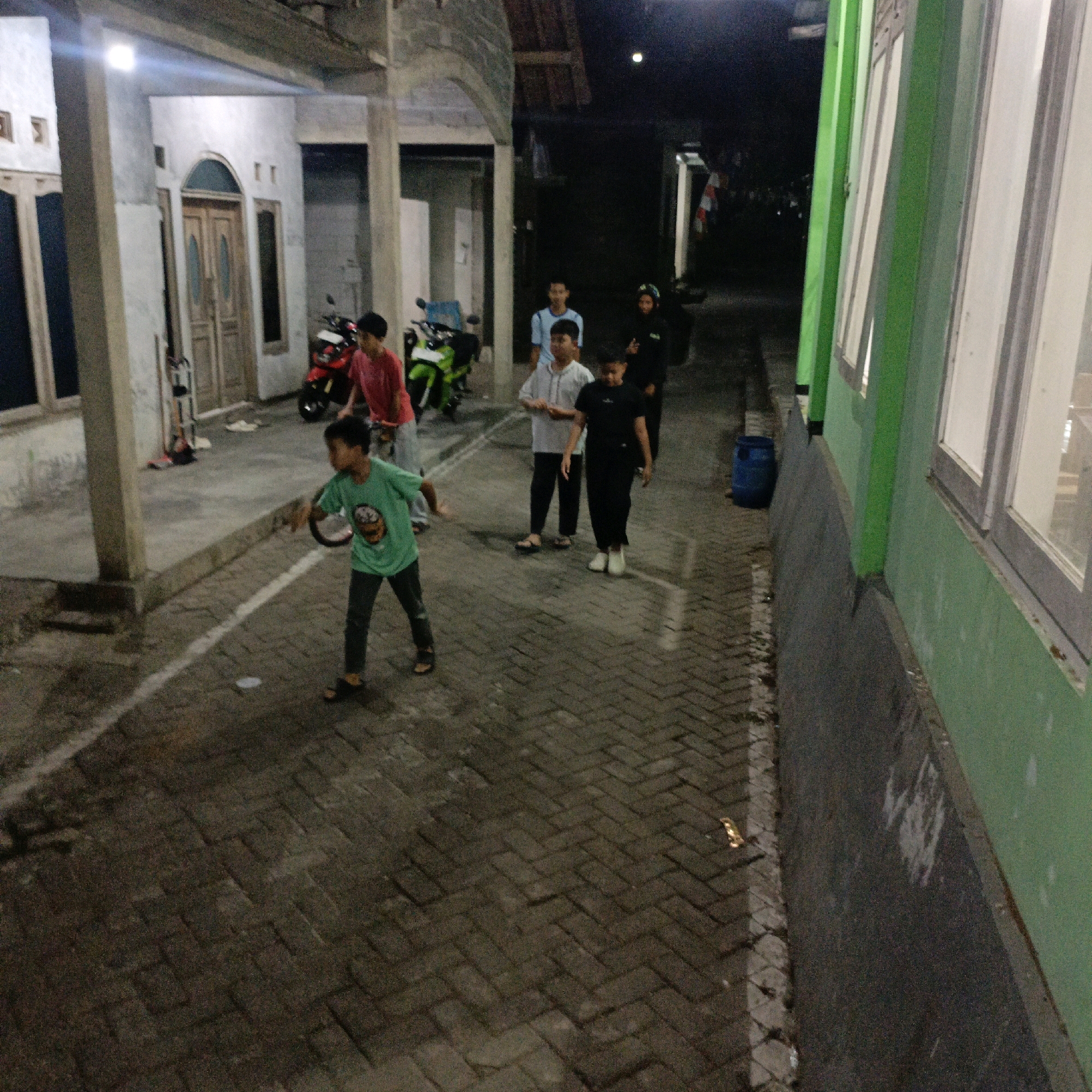 A group of young people are gathered on a paved walkway at night, near a building with green and white walls. In the foreground, a boy in a green t-shirt and dark pants is shown in mid-action, as if throwing something. To his left, a boy in a red t-shirt and dark pants is standing near a red motorcycle. Behind them, other individuals are visible, including a boy in a light blue t-shirt and another in a white shirt and dark pants, and a person in dark clothing. Further back, a green motorcycle is parked. The environment appears to be residential or a public space, possibly in Salatiga, Indonesia, characterized by the brick-patterned pavement and the building architecture. A blue barrel is situated to the right side of the frame.