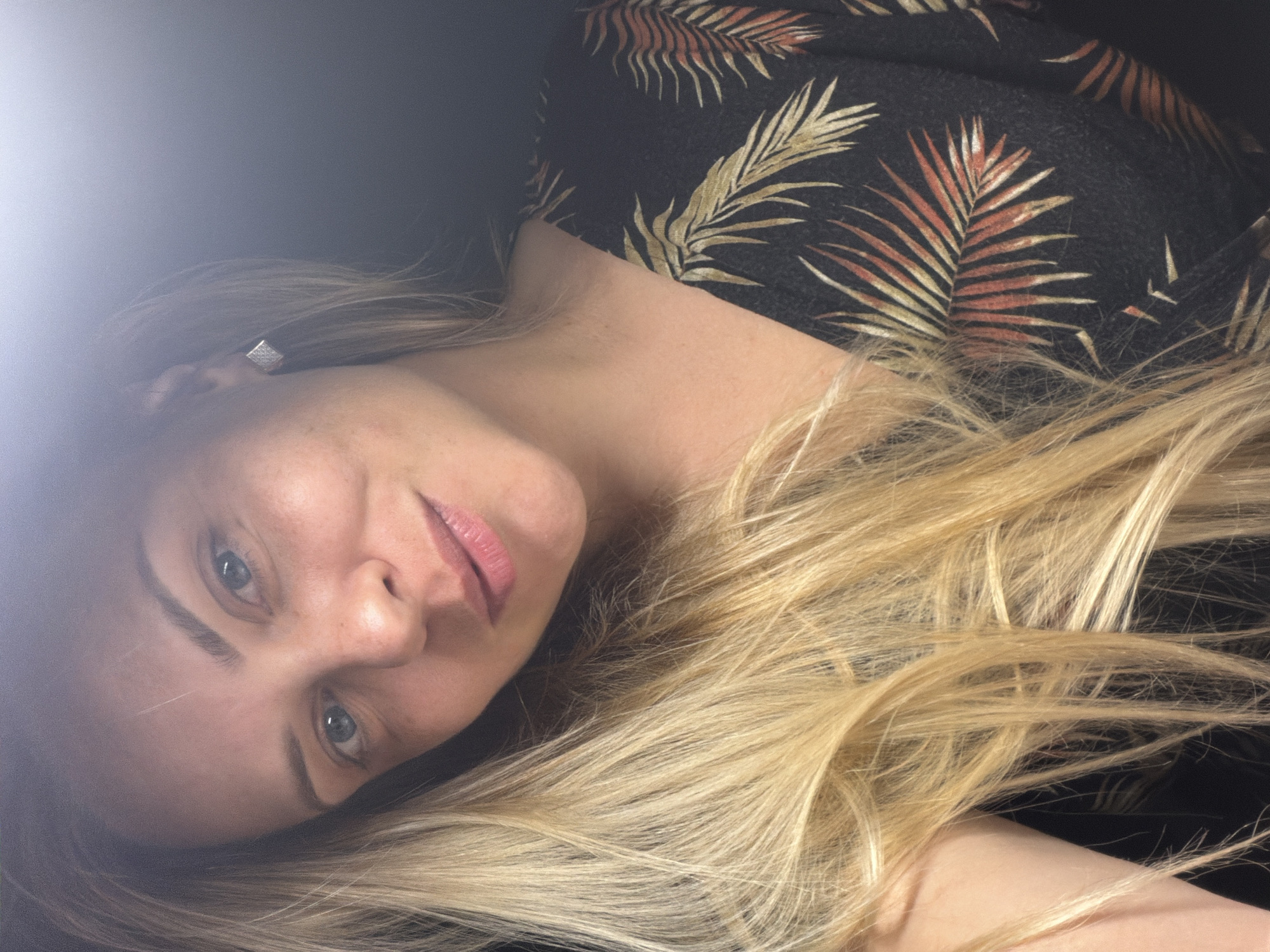 A close-up, overhead, shoulder-level shot shows a blonde woman posing for a selfie in Votkinsk, Russia. The woman has fair skin, light blue eyes, and fair skin. She is wearing a black dress with a tropical palm leaf print in shades of green, yellow, and orange. Her blonde hair is long and wavy, framing her face. She is wearing small, square stud earrings. The background is dark, with a bright light source on the upper left, creating a blurred effect and obscuring details of the environment.