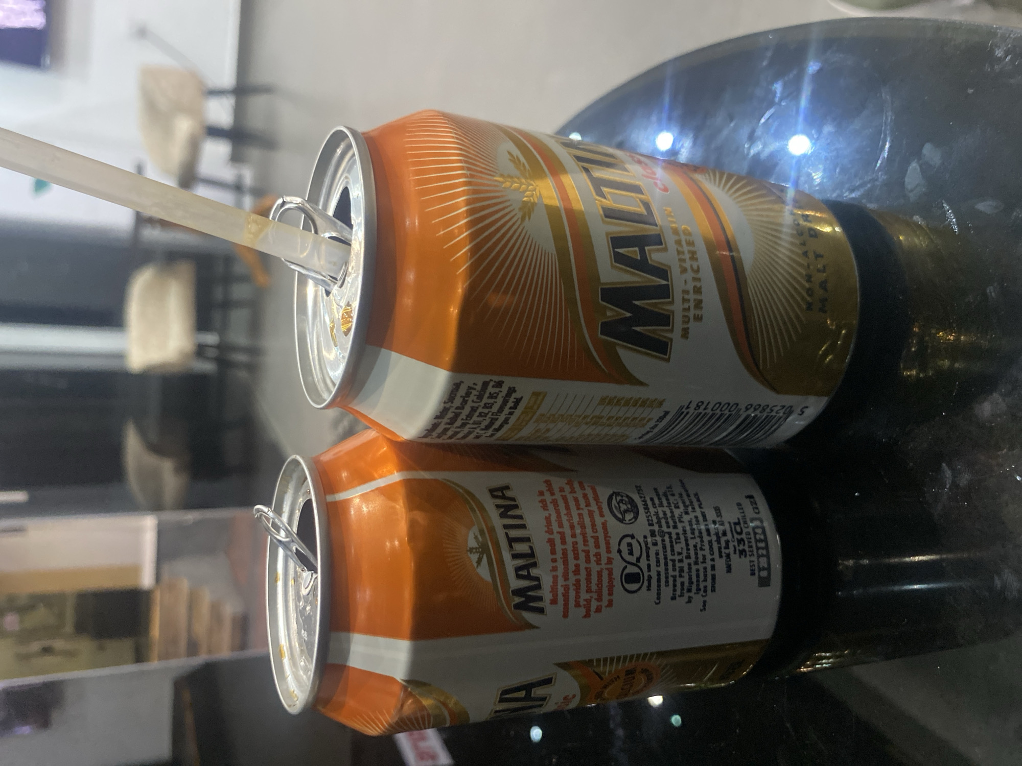 Two cans of Maltina, a non-alcoholic malt drink, are on a reflective black surface. The can on the left is partially visible and the can on the right is in focus. The right can is orange and white with "MALTINA" printed in large black letters, and "MULTI-VITAMIN ENRICHED" below. A straw is inserted into the opened top of the right can. The background is blurred, showing what appears to be an indoor setting with chairs and a doorway. The image is well-lit, with reflections on the surface. There are no people visible in the image. The location is identified as Barden Yana, Nigeria, based on the label on the can which mentions "Brewed and canned under license from PAI B.V., The Netherlands, by Nigerian Breweries Plc, P.C. 612, Igname House, Lagos, Nigeria."