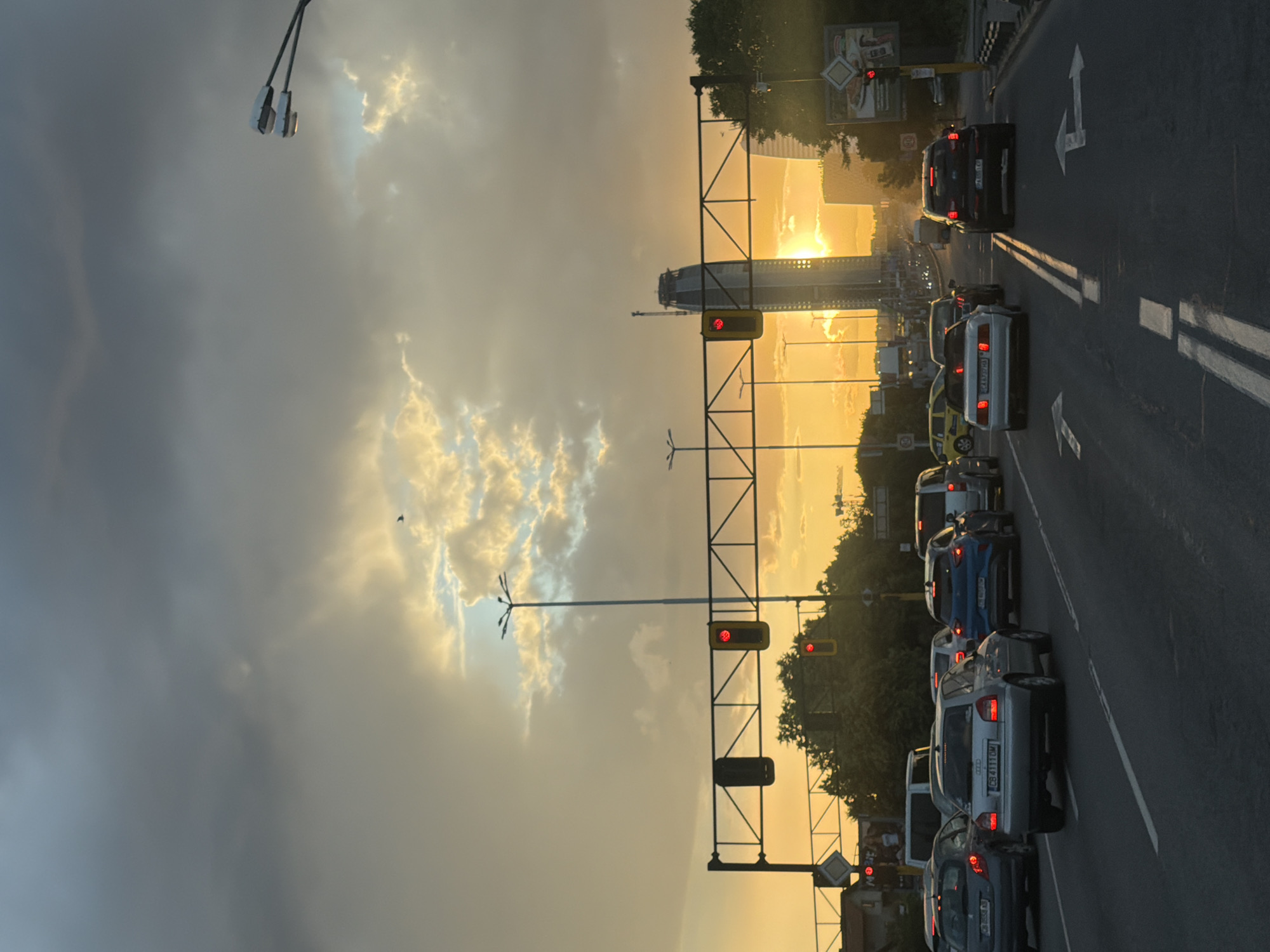 The image depicts a road scene in Sofia, Bulgaria, during sunset. The setting is a multi-lane road with cars, suggesting traffic. The sky is filled with dramatic clouds, illuminated by the golden light of the setting sun. The sun is low on the horizon, casting a warm glow over the scene and highlighting the textures of the clouds.
Several cars are visible on the road, indicating a traffic jam or slow-moving traffic. The cars are of various makes and models, and some have their brake lights on. Traffic lights are also visible, with at least one showing a red light, which is consistent with the traffic situation. In the background, there are buildings, including what appears to be a tall, modern structure, possibly a skyscraper or a distinctive landmark. Trees line the sides of the road, and there are streetlights and other infrastructure associated with a roadway. A bird is visible in the sky, adding a natural element to the urban scene.
The overall mood is one of a typical evening commute in a city, enhanced by the striking natural beauty of the sunset. The contrast between the urban environment and the dramatic sky creates a visually interesting composition. The presence of street signs and road markings further reinforces the location as a public road in an urban area. Some text is visible on license plates of the cars and on signs in the background, but it is not clear enough to read accurately.