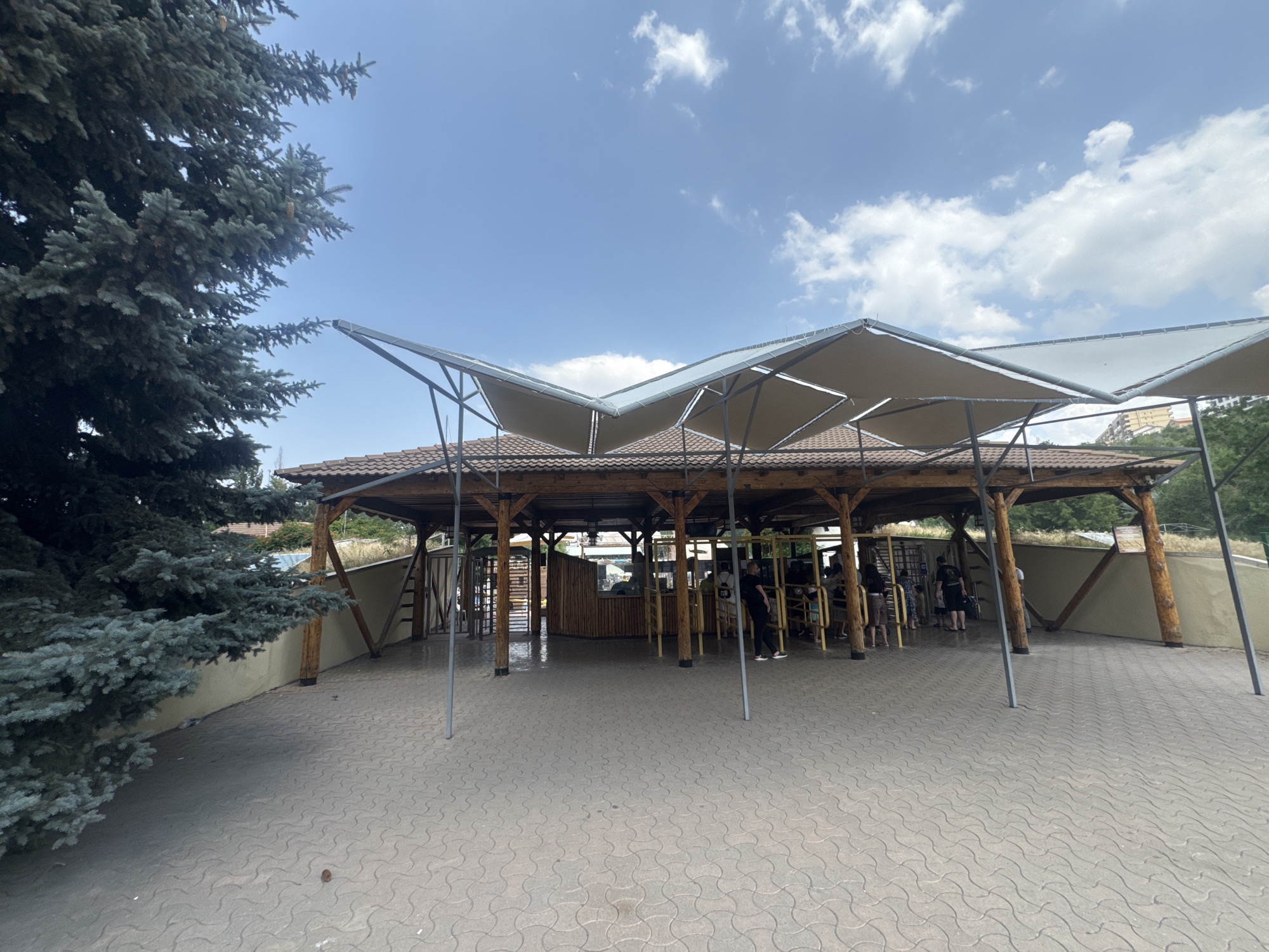 The image shows an outdoor area with a wooden structure providing shelter, likely an entrance or waiting area. The structure features a tiled roof and is supported by wooden beams. A modern canopy of fabric squares is stretched over the roof, creating a unique architectural element. In front of the structure, there are turnstiles, and several people are visible, some queuing and others walking. The ground is paved with interlocking concrete tiles. To the left, a large evergreen tree with blue-green needles dominates the foreground, its branches extending towards the sky. The background shows a building and lush greenery, suggesting a park or recreational area within a city. The sky is blue with scattered white clouds, indicating a pleasant day. Based on the context provided by similar images available online, this scene is identified as the entrance to the Yerevan Zoo in Yerevan, Armenia.