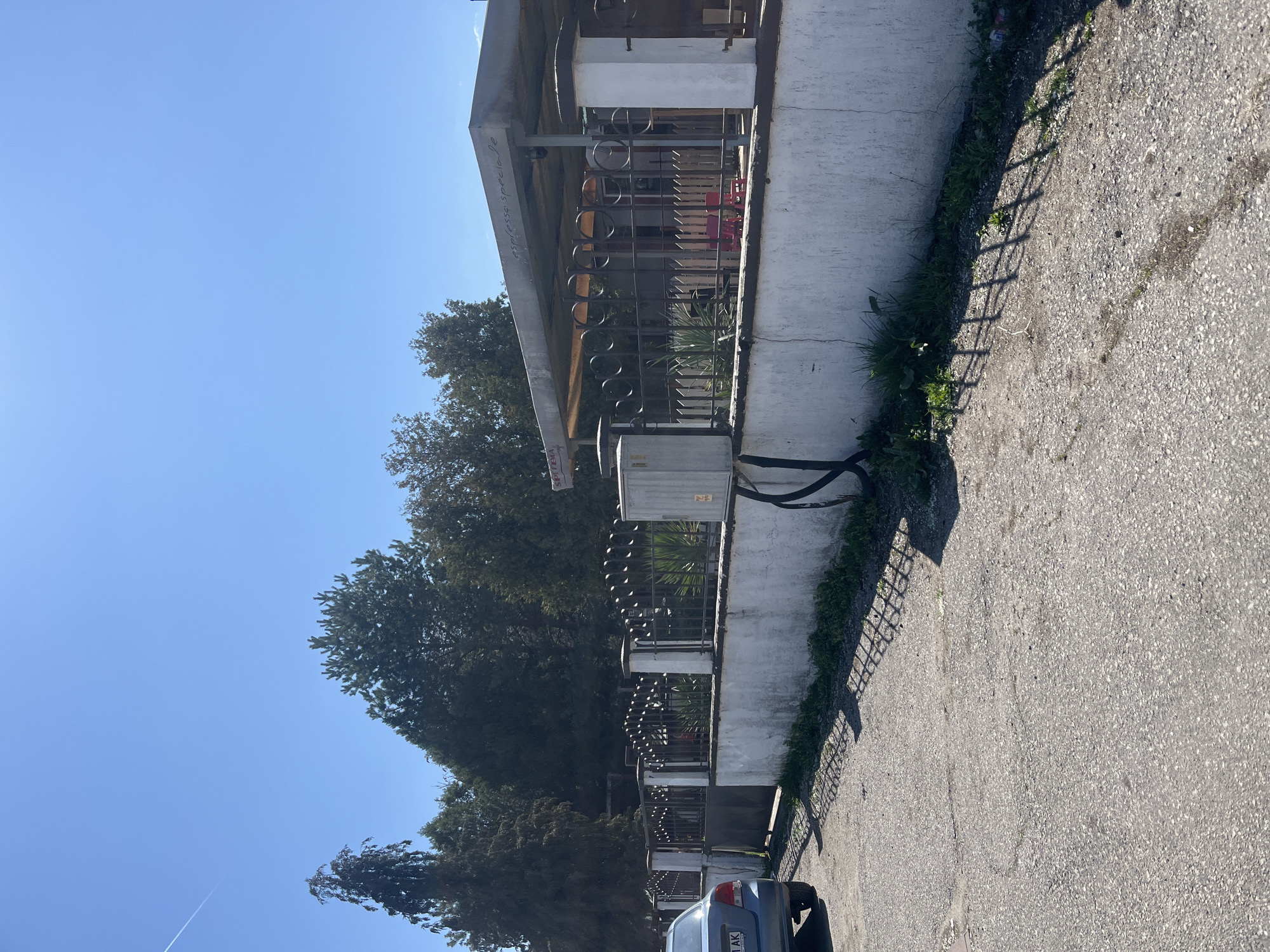 The image depicts a street scene in Sofia, Bulgaria. A building with a white facade is visible along with a metal fence. There is an electrical box attached to the building. Trees line the street. A blue car is parked on the right side of the street. The sky is clear and blue. The ground is paved with asphalt.