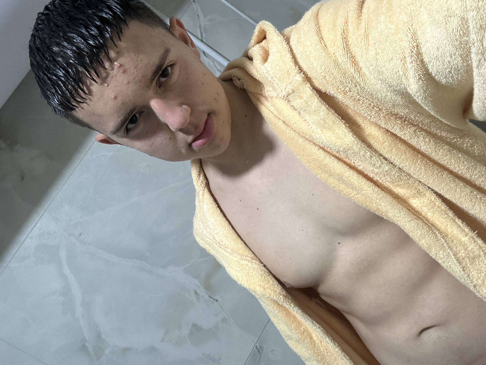 An eye-level selfie captures a young male indoors, likely in a bathroom or shower area. He has short, dark, wet hair with water droplets visible on his forehead and skin, suggesting he has recently showered or bathed. His face, angled slightly, shows dark eyebrows and eyes, with some acne or blemishes visible on his forehead and cheeks. He wears a light yellow or pale orange terry-cloth bathrobe or towel draped loosely around his shoulders, revealing his bare chest and upper abdomen, indicating a lean physique. The background consists of light grey or white tiled walls with subtle marbling patterns. The lighting is bright and even, consistent with an indoor setting. The subject's expression is neutral, looking directly at the camera. There is no visible text.
