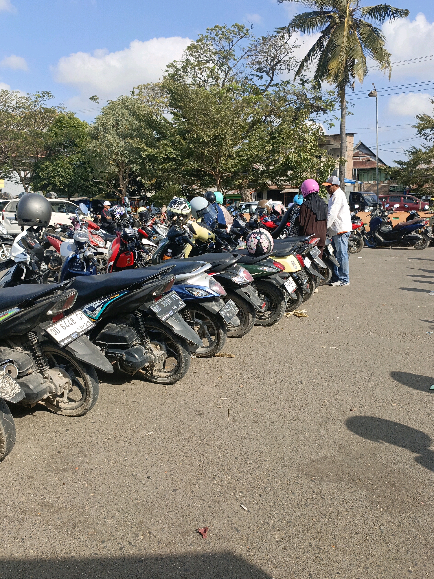 The image shows a parking lot filled with motorcycles, likely in the city of Bulukumba, Indonesia. The weather is sunny with some clouds. Several people are visible, some wearing helmets and others without. One person, a man in a white shirt and blue jeans, is standing next to a woman wearing a pink hijab and a brown abaya. The motorcycles are parked in rows, with some facing forward and others backward. The ground is asphalt, and there are trees and buildings in the background. The overall scene suggests a busy area where people are parking their vehicles, possibly for an event or to visit a nearby establishment.