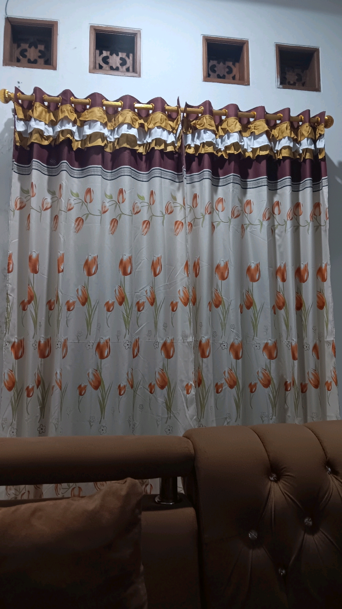 The image depicts a close-up view of a window with a decorative curtain featuring a floral pattern of orange tulips. Above the curtain, three small, square-shaped wooden window vents are visible, each with an ornate carved design. The wall appears to be painted white. In the foreground, a portion of a brown tufted sofa is visible, with a pillow resting on it. The scene is indoors, and based on the style of the windows and the overall aesthetic, it could be located in a residential setting in Depok, Indonesia. No people are present in the image.