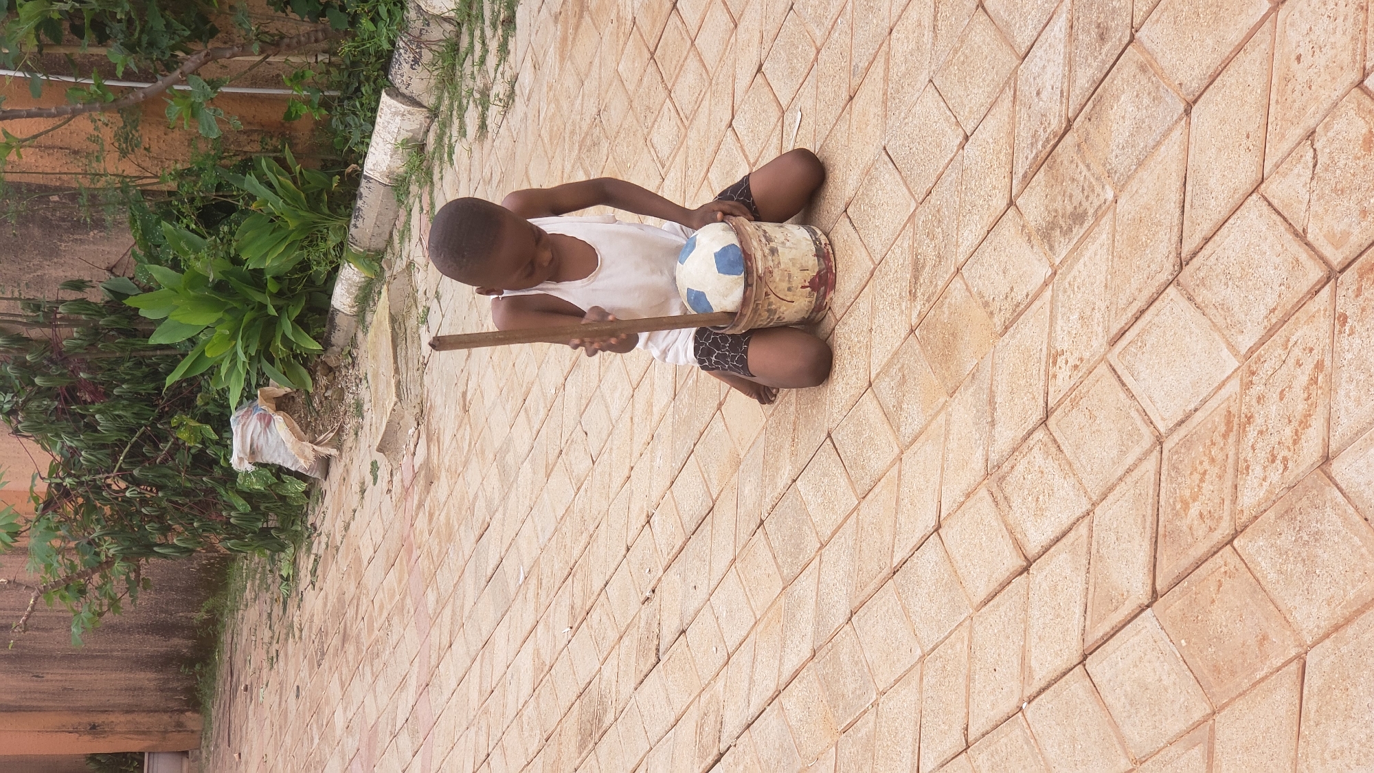A young boy kneels on a paved courtyard in Sabon Tasha, Nigeria, holding a wooden stick over a bucket containing a soccer ball. The boy, wearing a white tank top and patterned shorts, is focused on the bucket and ball. The courtyard is paved with rectangular terracotta-colored tiles laid in a diamond pattern. In the background, lush green foliage grows against a textured, light orange wall. A plastic bag filled with debris rests on the ground near the wall. The scene appears to be outdoors, in a residential or public space.