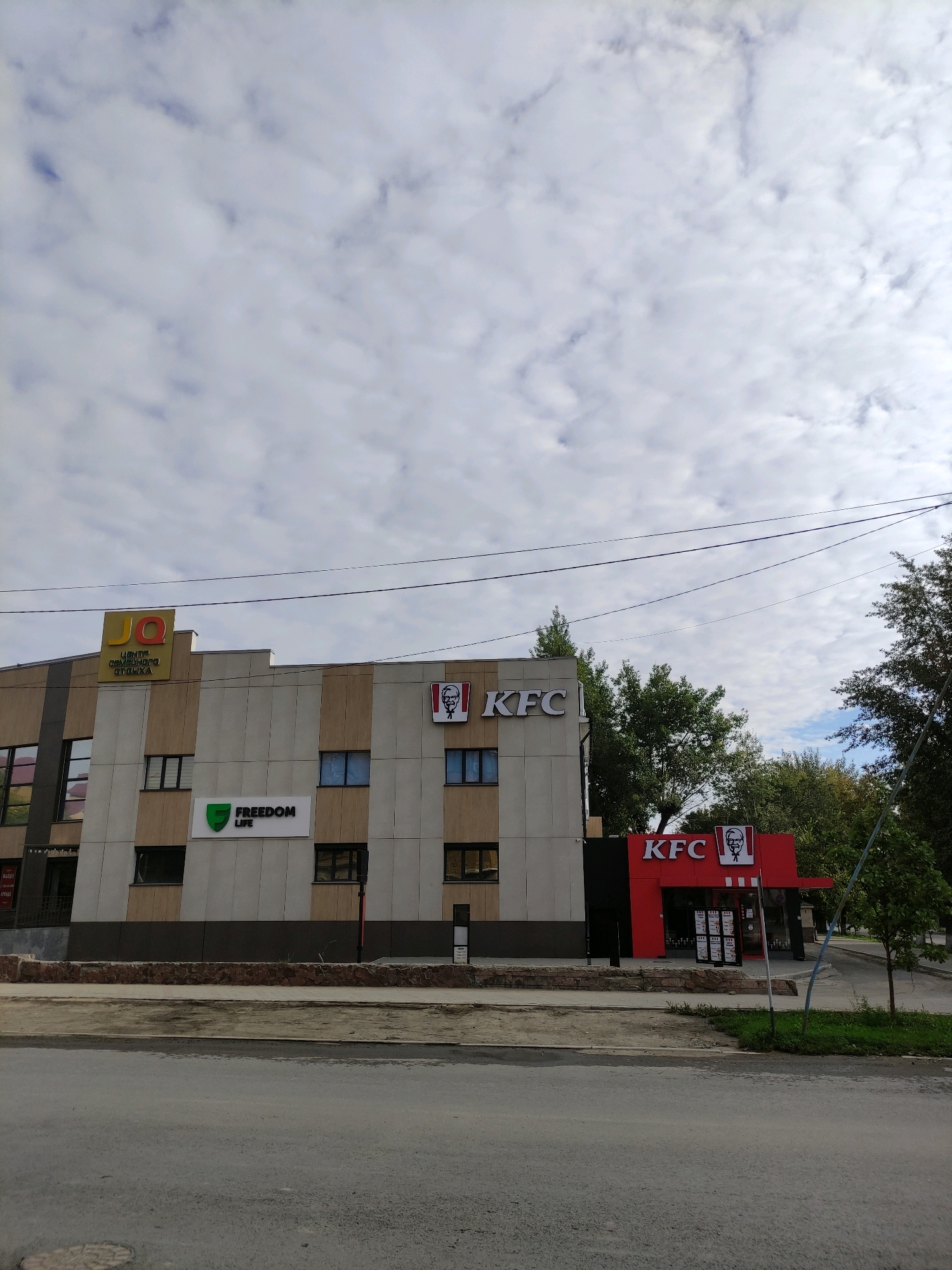 The image shows a two-story building with multiple businesses. The building is light-colored with vertical stripes of beige and brown. On the upper part of the building, there is a yellow sign with the letters "JQ" and Russian text below it, which translates to "Center for Family Leisure." To the right of this sign, also on the upper level, are two KFC logos. On the lower level, there is a sign that reads "FREEDOM LIFE." Further to the right, there is a red KFC restaurant with its distinctive logo and menu boards visible through the windows. The building is located on a street in Semey, Kazakhstan, as indicated by the context. The street in front of the building appears to be paved, with a sidewalk and a low stone wall separating it from the building. Trees and greenery are visible behind the building. The sky is overcast with white and gray clouds. There are no people or vehicles clearly visible in the frame.