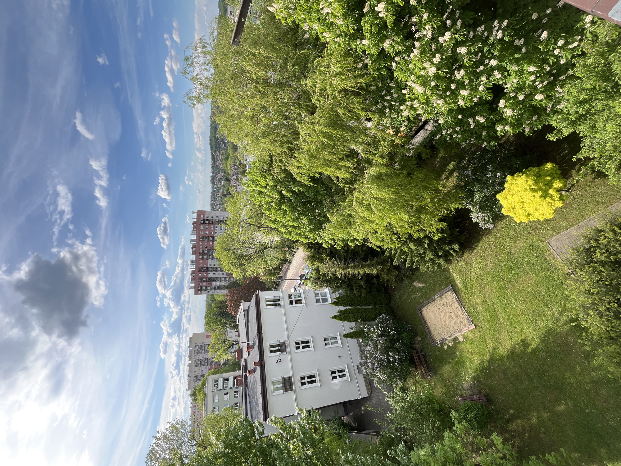A high-angle view presents a cityscape on a sunny day, under a blue sky dotted with white clouds. In the foreground, lush green trees and a patch of grass dominate, with a small sandbox visible on the right. Adjacent to the trees are residential buildings, painted in light hues with visible windows and roofs. Beyond the buildings, taller apartment complexes stand in the background. The location appears to be a residential area, characterized by a mix of green spaces and urban infrastructure.