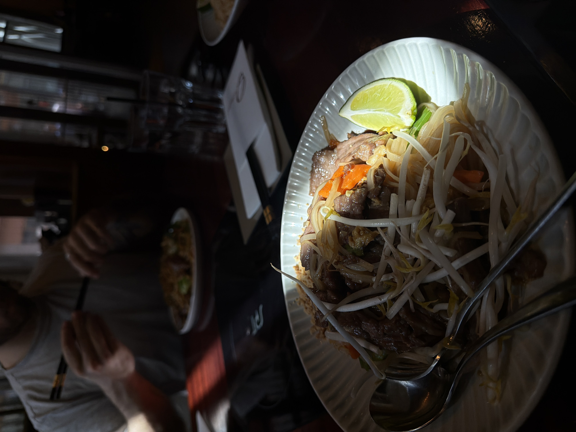 The image shows a plate of Pad Thai with a lime wedge, set on a dark surface. A spoon and fork are visible on top of the noodles. There are other dishes and a person holding chopsticks in the blurred background. The light source is casting a strong shadow and highlight on the plate. Without further information, it is impossible to confirm its exact location in Boston or even if it is Boston at all.