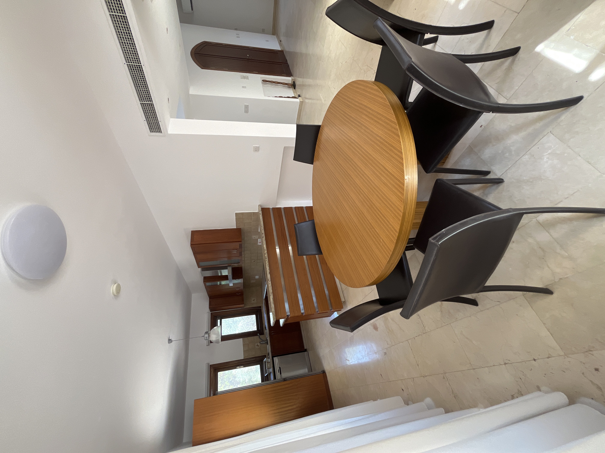 This image depicts an indoor scene, likely a dining area adjacent to a kitchen, within a residential property located in Aphrodite Hills, Cyprus. The room features a light-colored tiled floor and white walls. In the foreground, a circular wooden dining table is set with six black chairs. The table has a light wood grain pattern. Behind the dining area, a kitchen is visible, with dark wood cabinetry, a granite countertop, and stainless steel appliances. A breakfast bar with horizontal wooden slatted panels separates the kitchen from the dining area. Large white curtains are visible on the left side of the image, partially obscuring a tall wooden cabinet. In the background, a doorway with a curved wooden top is visible, leading to another area of the property. The overall impression is of a modern, well-lit residential interior.