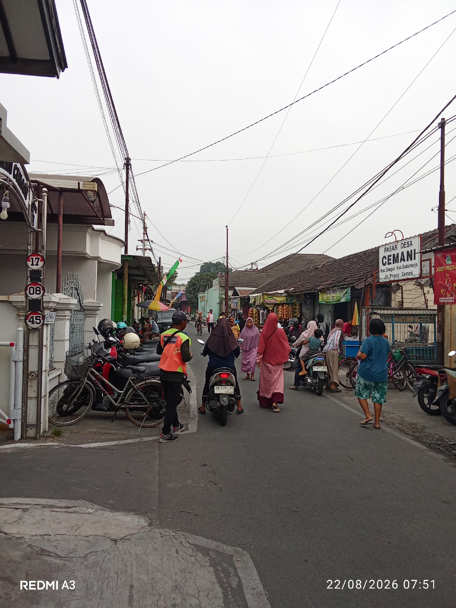 The image depicts a street scene in Cemani, Indonesia, with a market visible in the background. A variety of people are present, some walking and others riding motorcycles. Several motorcycles are parked on the side of the road, along with a bicycle. Overhead, numerous power lines crisscross the sky. Building facades with awnings line the street, suggesting commercial activity. Traffic signs are visible, including one with the numbers "17," "08," and "45." The overall atmosphere appears to be that of a busy, everyday street in a local Indonesian community.
