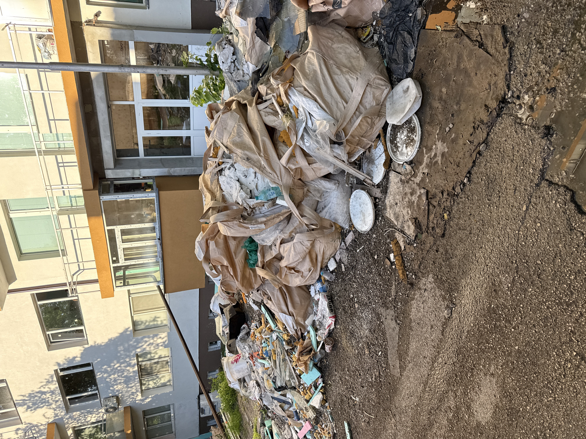 The image shows a large pile of construction debris and trash dumped next to a multi-story building. The building has white walls and windows, with some brown and tan accents. The debris includes large beige bags, pieces of insulation foam, broken plastic, and empty paint containers. The ground is cracked asphalt and dirt. The exact location in Lovech, Bulgaria cannot be determined from the image alone. There are no people visible in the image.