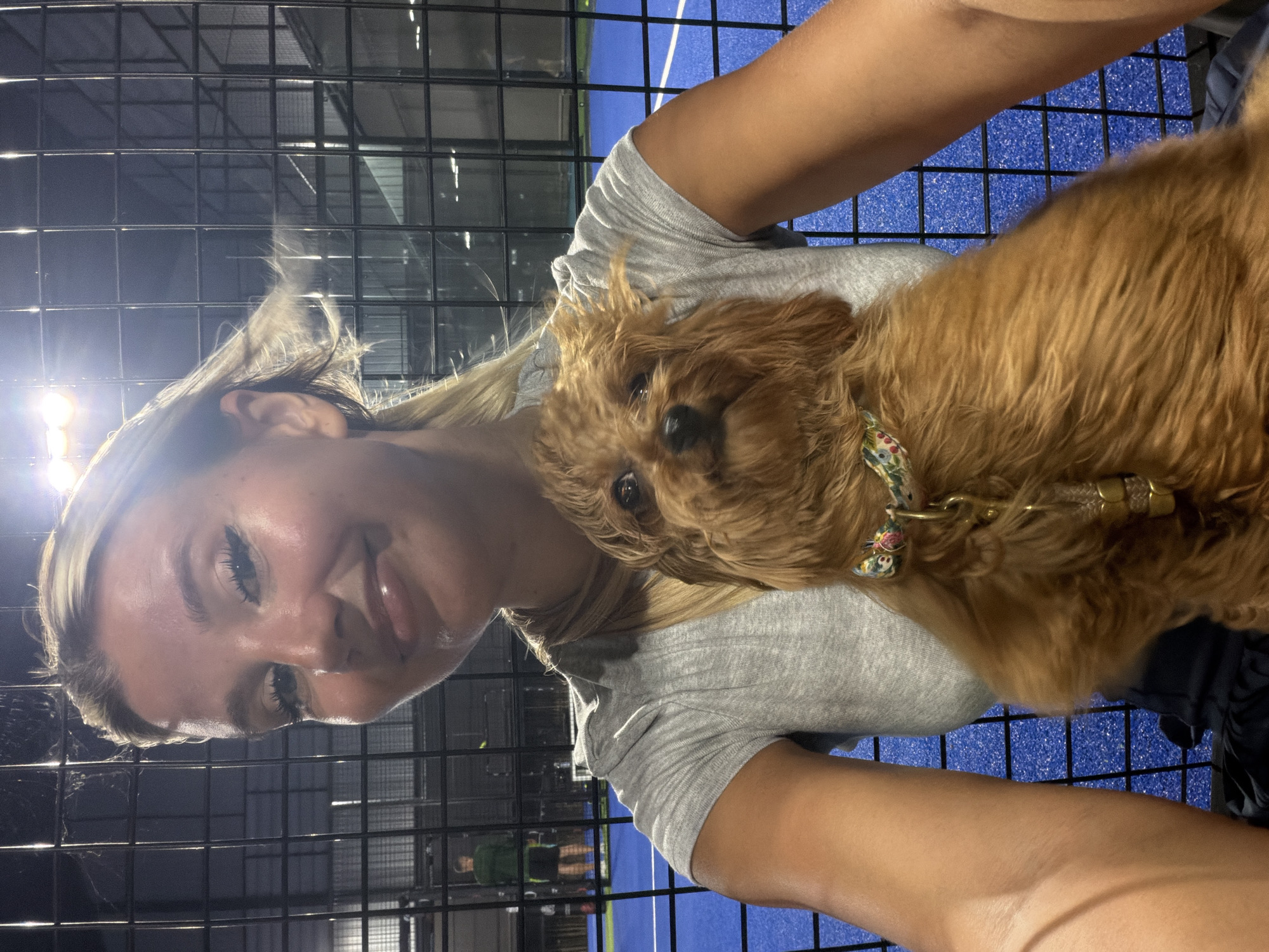 A woman and a dog are in a photo at a Padel tennis court. The woman has blonde hair and is wearing a grey t-shirt. She is holding a small, tan-colored dog with curly fur. The dog is wearing a collar with a floral pattern. The background shows a blue Padel court with black fencing. There are bright lights illuminating the court from above. In the background, a man in a green shirt and black shorts is visible on the court. The woman is looking at the camera and smiling. The dog is looking at the camera.