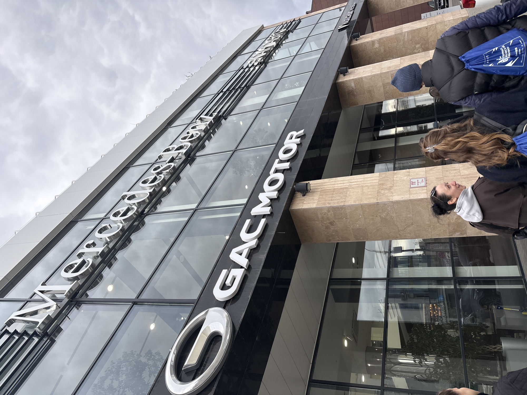 The image depicts a modern building with a glass facade in Kazan, Russia, identified as a GAC Motor dealership. The GAC Motor logo and name are prominently displayed on the building's exterior. Several people are standing on the sidewalk in front of the building, including a woman with sunglasses on her head wearing a brown jacket with white hood, a person with long brown hair and a silver clip wearing a blue string bag, and a person wearing a blue hat and a black jacket. The sky is overcast, and reflections of the sky and surroundings are visible in the glass facade.