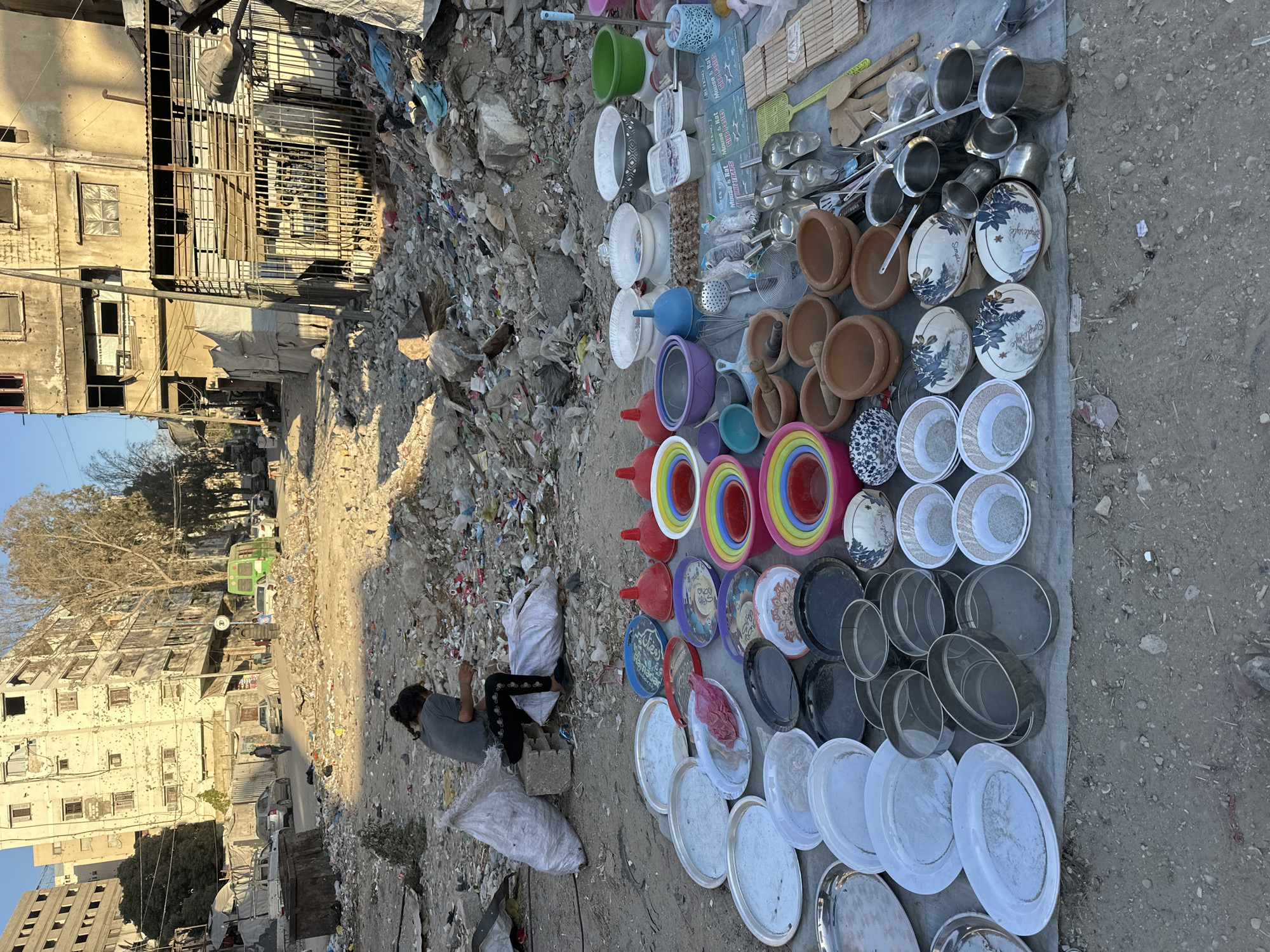 A person sits in the foreground on the left, hunched over sorting through debris. They are next to two large white sacks, one bulging. In front of the person, laid out on a tarp, is a collection of various household items, mostly kitchenware. These include several large, shallow metal platters, a stack of nesting colorful plastic bowls, earthenware bowls, metal sieves, and various utensils. Behind the person and the tarp is a street scene littered with rubble and debris. Several damaged buildings line the street, some appearing partially destroyed. A few vehicles are visible, including a green van. The overall environment is one of urban decay and apparent poverty, with goods being sold or sorted amidst the ruins.