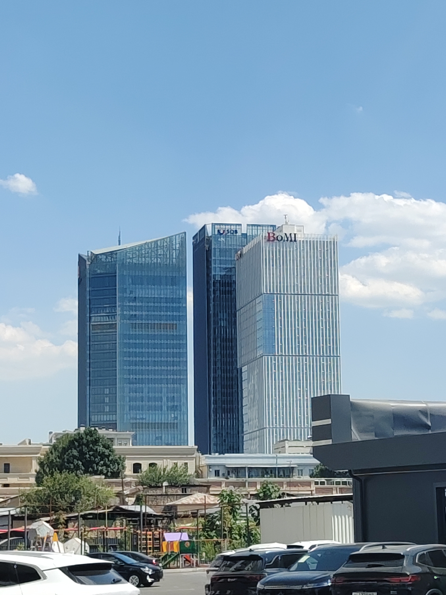 In a daytime outdoor scene in Tashkent, Uzbekistan, three modern skyscrapers dominate the background under a bright blue sky with scattered white clouds. The leftmost skyscraper is tall and slender with a predominantly glass facade, featuring a distinct angled top. To its right stands another equally tall skyscraper with a darker glass exterior. The third skyscraper, also tall and featuring a glass facade, is located to the right of the previous two and displays the red lettering "BoMI" prominently at its apex. In the mid-ground, a row of lower buildings with yellow and beige facades is visible, interspersed with lush green trees. The foreground shows a dark grey structure with a flat roof, partially obscuring a line of parked cars. Among the cars are several black SUVs and a white SUV, all facing away from the viewer. A children's playground with colorful equipment is visible in the lower right quadrant of the mid-ground, behind a section of fencing.