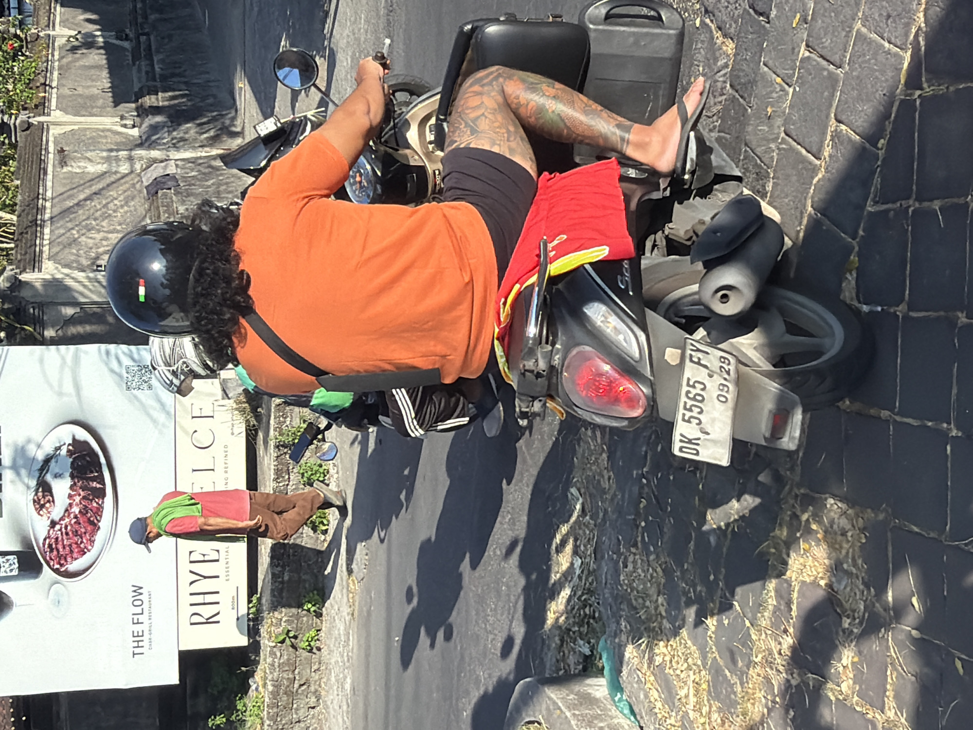 In the city of Mengwi, Indonesia, on a sunlit, paved road, a man is riding a scooter. He has long, dark, curly hair and wears a black helmet with an Italian flag emblem. He is dressed in an orange t-shirt and dark shorts, and his legs are heavily tattooed. A red cloth is draped over the back of the scooter. To the left of the scooter, a man in a red shirt, brown pants, and a blue cap is walking away from the camera. In the background, a large billboard advertises "THE FLOW CHAR-GRILL RESTAURANT" and "RHYE ELCE ESSENTIAL". The environment appears to be a street scene with some foliage and stone structures visible.