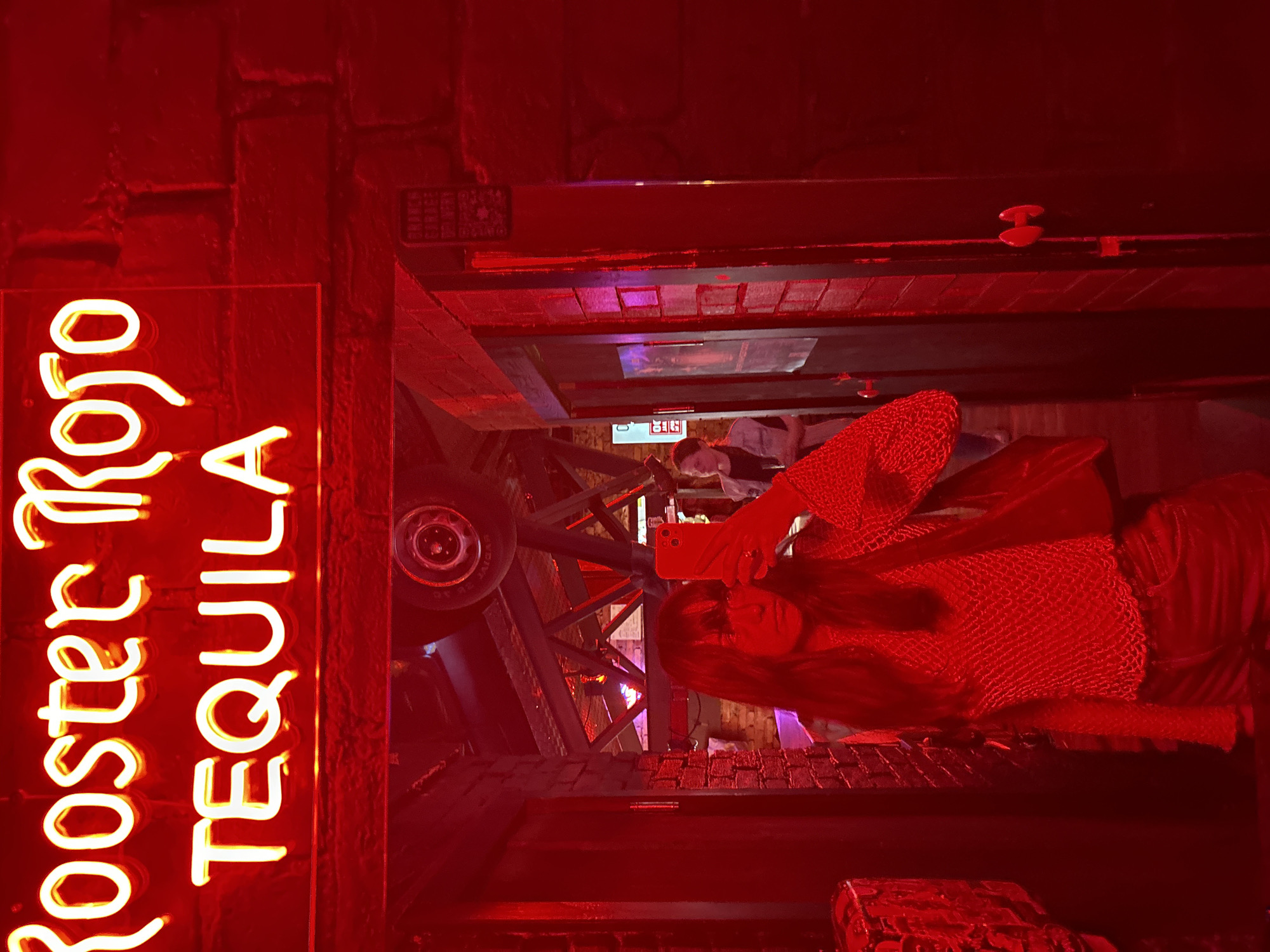 The image displays an interior scene, characterized by a pervasive red light, likely a bar or club in Saint Petersburg, Russia. Dominating the upper portion is a neon sign that reads "Rooster Rojo" in script above "TEQUILA" in block letters, illuminated in bright yellow-white against a red brick wall.
In the foreground, an individual with long, dark hair and bangs is visible in a mirror reflection, capturing a selfie with a red or orange smartphone. This person is wearing a red, open-weave mesh top over a darker garment, possibly paired with dark shorts or jeans, and has a ring on their finger.
The reflected background reveals an environment constructed with red brick walls and dark wooden beams or paneling. Suspended from the ceiling structure, which includes metal trusses, is a wheel-like object, possibly a car wheel or tire, serving as decor. Further back in the venue, another person, appearing to be female with lighter hair, stands near a doorway, wearing a light-colored vest over a dark top. Several dark doorways are visible, one of which has a small poster and another features distinctive red horizontal handles. A dark rectangular sign with partially discernible white text, including the phrase "STAFF ONLY," is affixed to a wooden frame to the right of the primary reflection.