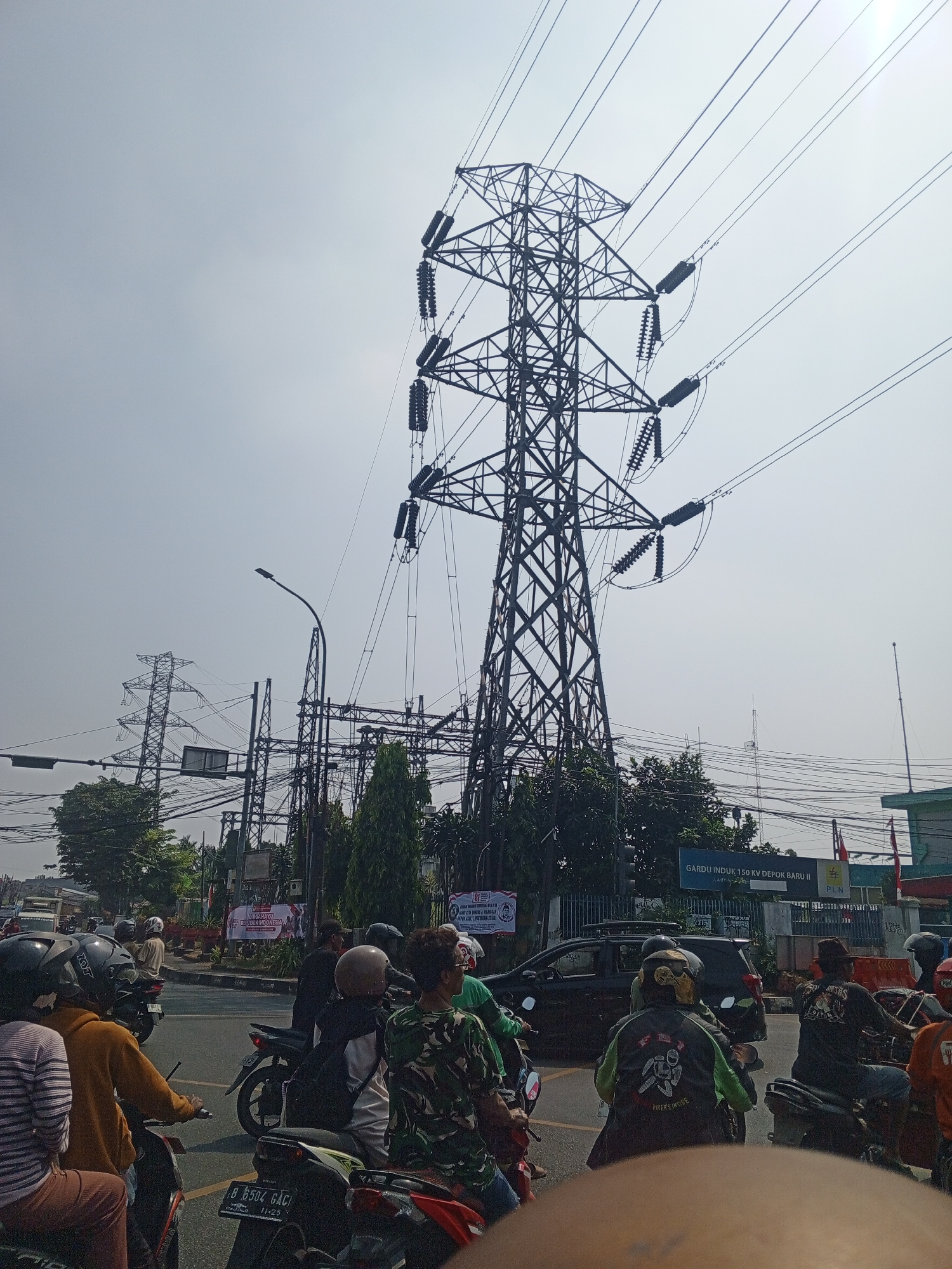In Depok, Indonesia, a busy street scene unfolds under a hazy sky, dominated by a towering electricity pylon. Several people on motorcycles are stopped at an intersection, awaiting the signal to proceed. The pylon, a lattice structure of steel, supports multiple high-voltage power lines that stretch across the frame. In the background, more electrical infrastructure, including substations and additional pylons, are visible, along with trees and buildings. A sign reads "GARDU INDUK 150 KV DEPOK BARU II" indicating the location as a 150 kV main substation in Depok. The overall atmosphere suggests a typical urban environment with significant power transmission infrastructure.