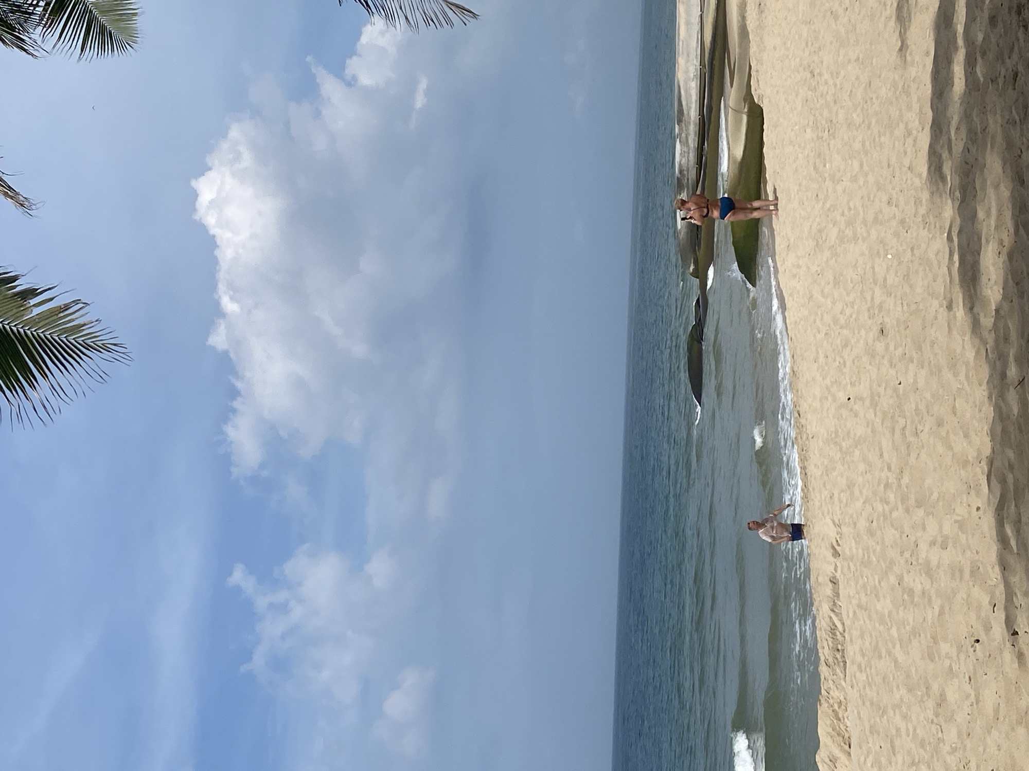 The image depicts a beach scene in Phu Thuy Ward, Vietnam. A sandy beach occupies the foreground, with palm fronds visible at the top corners. The ocean stretches out in the midground, with gentle waves lapping the shore. In the water, a man wearing a light-colored shirt and dark shorts is partially submerged, appearing to walk out of the sea. On the shore, near some large rocks and concrete structures, a woman in a blue bikini is standing and holding a phone, seemingly taking a photo. The sky is partly cloudy with blue sky visible.