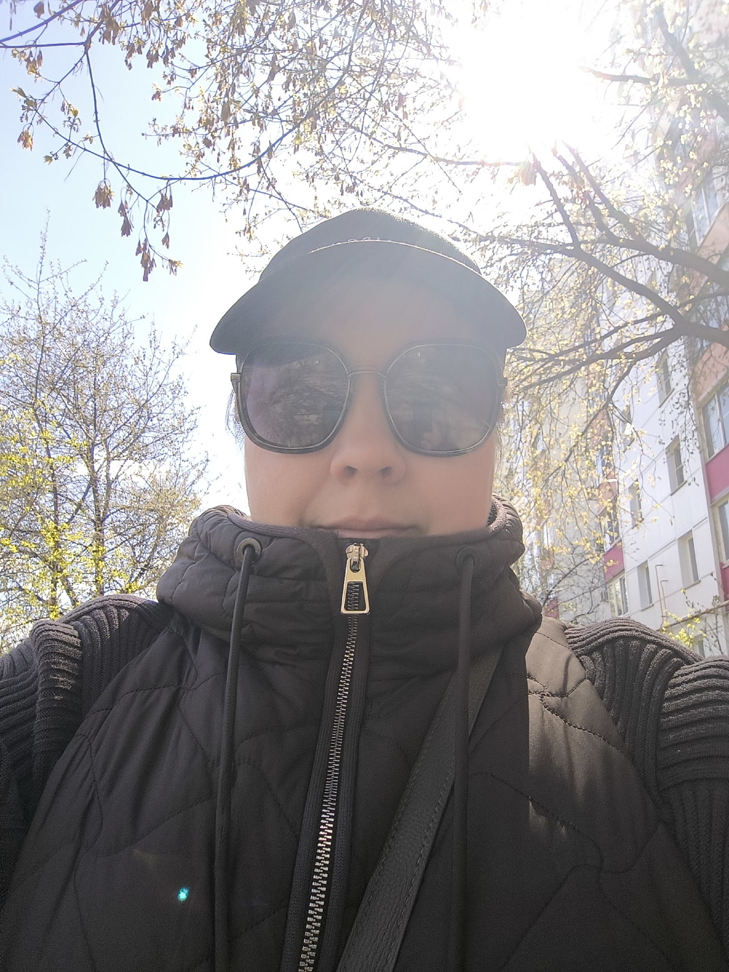 A selfie shot from a low angle captures a person wearing sunglasses and a black cap. The person is wearing a black quilted vest over a ribbed sweater or jacket, with the zipper of the vest visible. The background reveals a cityscape with trees and a building. The building is white with pink accents, and the trees are beginning to blossom, indicating springtime. The location appears to be a city, but the specific location remains unidentified.