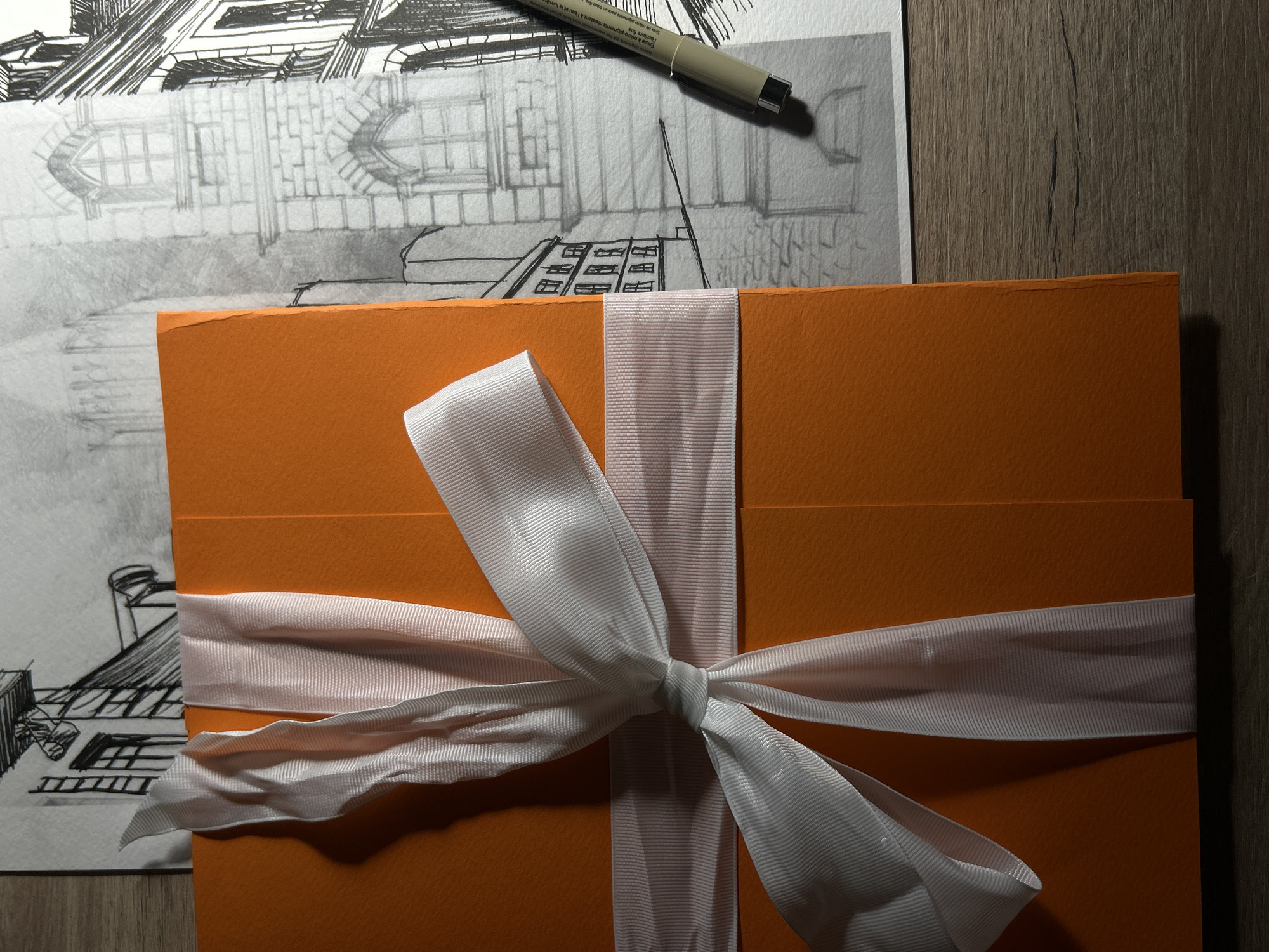 The image presents a top-down view of a creative workspace featuring an architectural drawing, a fineliner pen, and a ribbon-tied orange object, all arranged on a wooden surface.
On the left, a white sheet of paper displays an intricate architectural sketch. The drawing, rendered with both pencil and black ink, depicts a building facade with distinct arched windows, brickwork patterns, and roof structures, suggesting an urban or historical building. A black fineliner pen rests diagonally across the upper part of the paper.
Dominating the right and center of the frame is a stack of vibrant orange-colored paper or cardstock, which appears to be textured. This item is elegantly wrapped with a white, semi-transparent, ribbed ribbon, tied into a prominent bow in the middle. The ribbon casts soft shadows, indicating a focused light source.
The background is a dark, wood-grain surface, likely a desk or table, contributing to a professional and artistic setting.
The text visible on the fineliner pen's barrel, as per OCR, reads "Encre à micro-pigments résistant à l'eau et la lumi" (Ink with micro-pigments resistant to water and light), "l'écriture fine" (fine writing), and "pigmento para un trazo fin" (pigment for a fine stroke), highlighting the pen's durable and precise qualities.
While no direct activity is captured, the arrangement suggests an ongoing or recently completed artistic or design project, possibly preparing a gift or presentation. The photograph was taken in Sofia, Bulgaria.